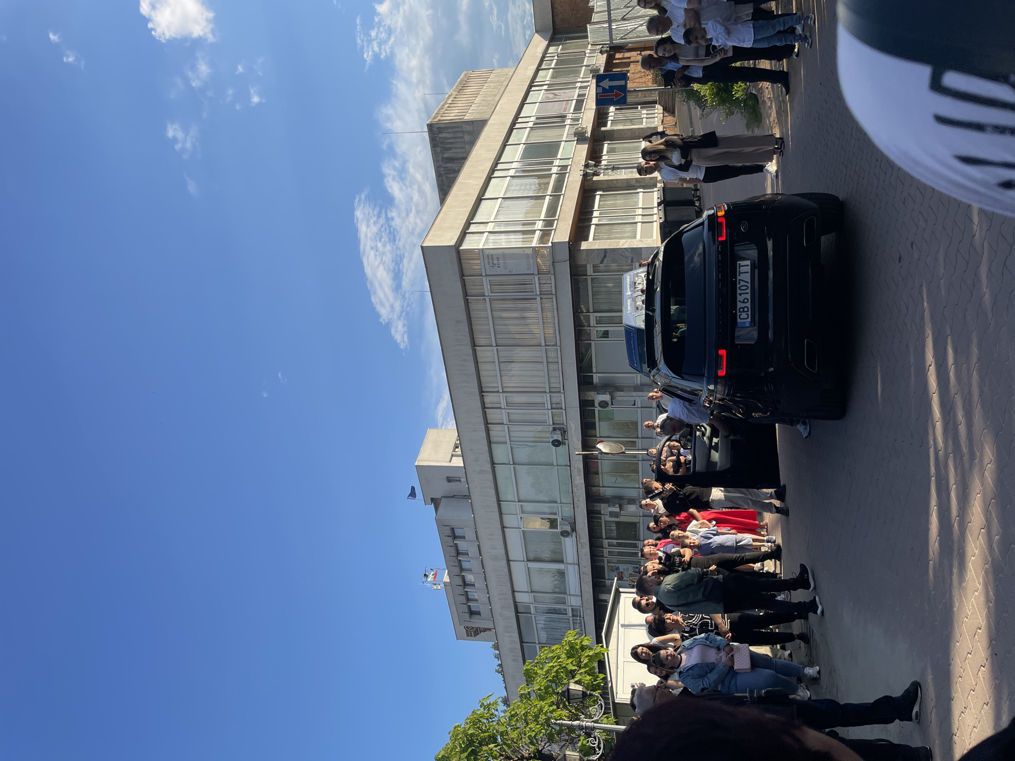 The image captures a scene in Zlatograd, Bulgaria, with a group of people gathered on a paved street near a large, multi-story building. A black Range Rover is visible, with its rear facing the camera and license plate "CB 6107 TT" displayed. People are standing along the street, and some are behind the car. The building has a modern facade with large windows, and flags are flown on its roof. The sky is blue with some clouds.