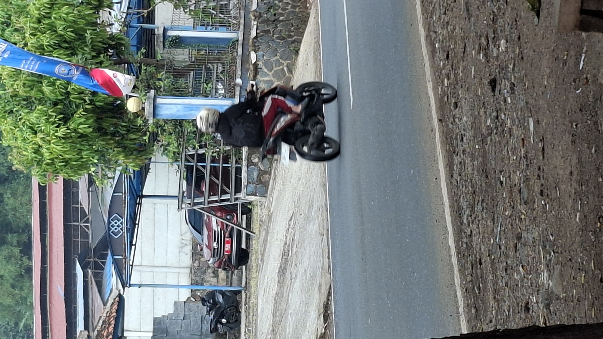 In Cilaku, Indonesia, a person wearing a black jacket and red pants rides a red and black motorcycle on a paved road. The rider is wearing a light-colored helmet and a black backpack. The motorcycle is in motion, with its wheels blurred. The scene is outdoors, with lush green trees in the background. A maroon SUV is parked on the side of the road near a building with a blue awning and a white wall. Another motorcycle is parked in front of the building. The area appears to be semi-urban, with elements of both natural landscape and man-made structures. A flag with red and white stripes is visible in the upper right portion of the image. The road surface is asphalt, with a white line marking the edge. The ground on the side of the road is covered with gravel and debris.