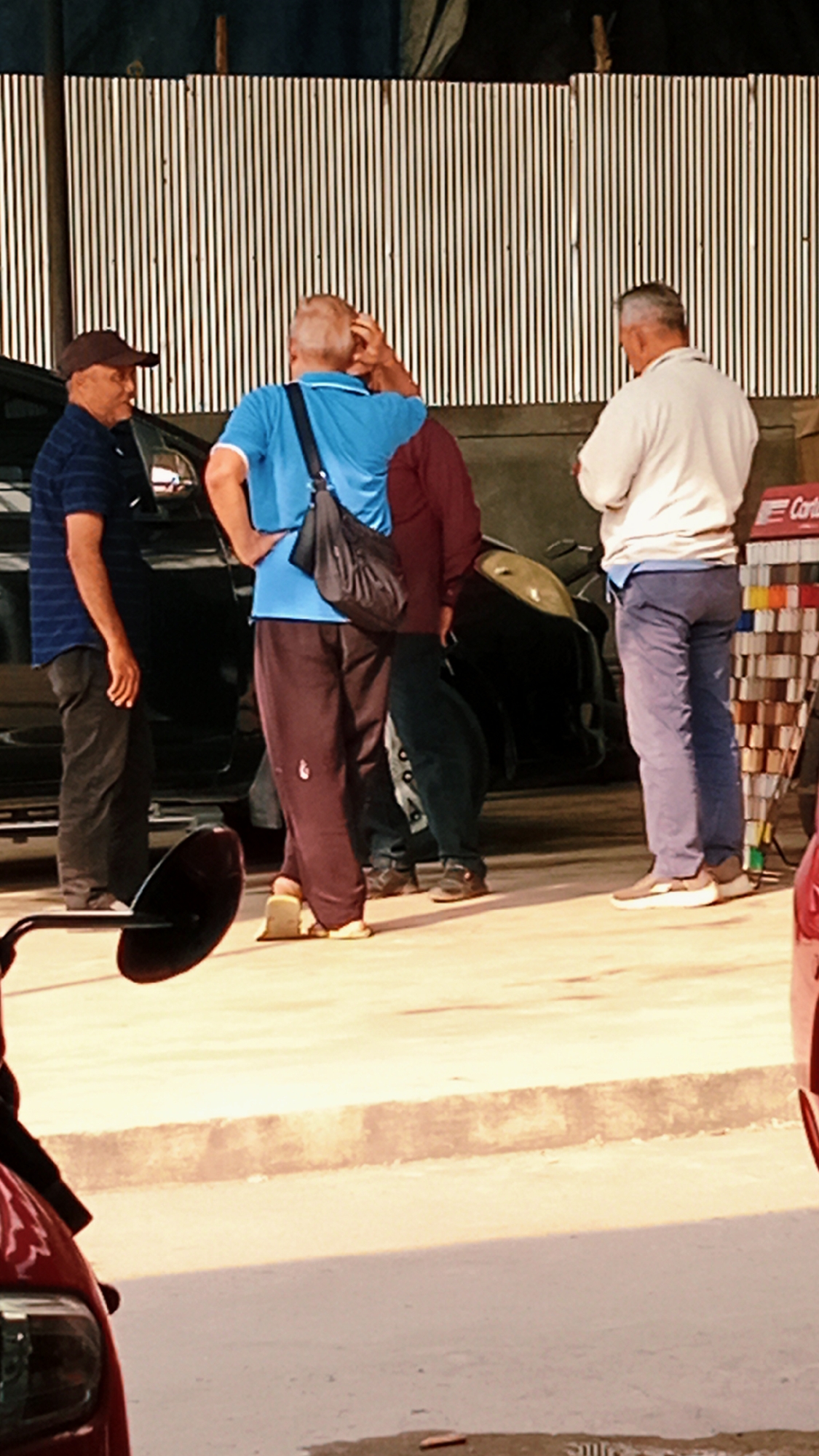 Four individuals are gathered outdoors in what appears to be a covered area, possibly a workshop or garage, with a corrugated metal wall in the background. The ground is paved with concrete.
On the left, a man wearing a dark baseball cap and a blue striped short-sleeved shirt stands facing right. He has his left hand on his hip. Next to him, a man wearing a blue short-sleeved shirt with a black crossbody bag slung over his right shoulder stands facing the center. His right hand is on his hip, and his left hand is near his head. Between these two men, partially obscured, is another individual wearing a dark reddish-brown jacket.
On the right, a man in a light-colored, possibly beige or off-white, long-sleeved shirt and blue jeans stands facing left, with his back to the camera. He appears to be looking down. Next to him, to the far right, is a display rack with colored items, possibly paint samples or tiles, and a red sign with white lettering that reads "Cort..."
In the foreground, a portion of a red vehicle is visible on the far right, and a black circular object, possibly a mirror or part of a vehicle, is on the left. The overall lighting suggests it is daytime.