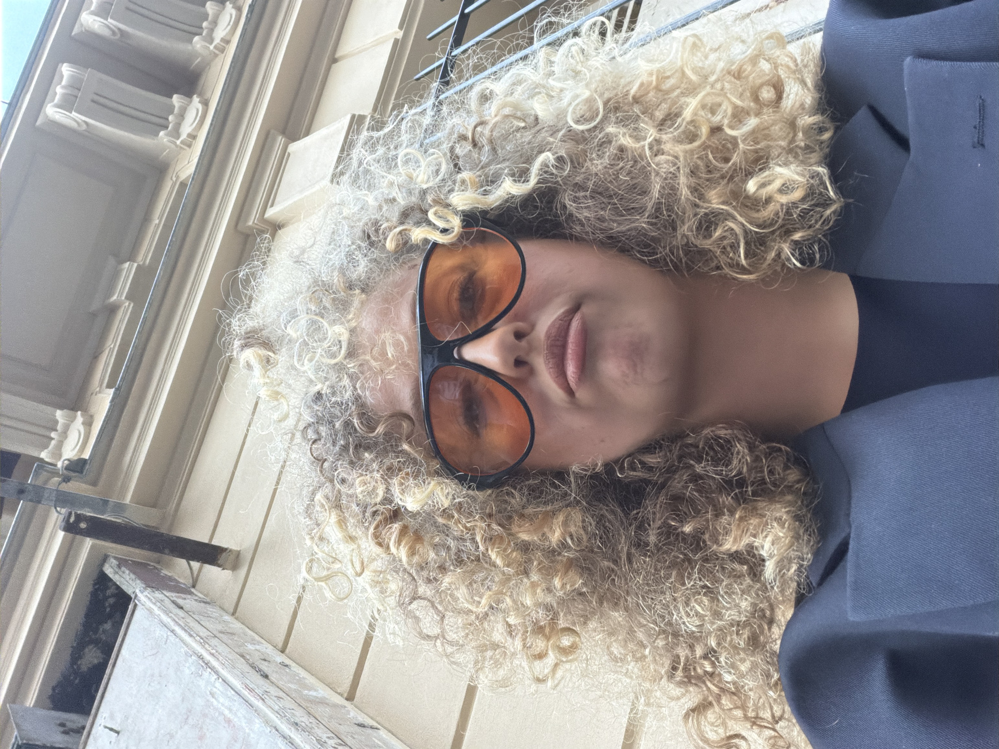 A woman with curly blonde hair and wearing black-rimmed sunglasses with orange lenses is posing in an outdoor setting. She is wearing a navy blue blazer. The background consists of a beige building with ornate architectural details and a metal grate. The location appears to be in Paris, France, based on the architectural style. The camera angle is looking up at the subject, slightly tilted.