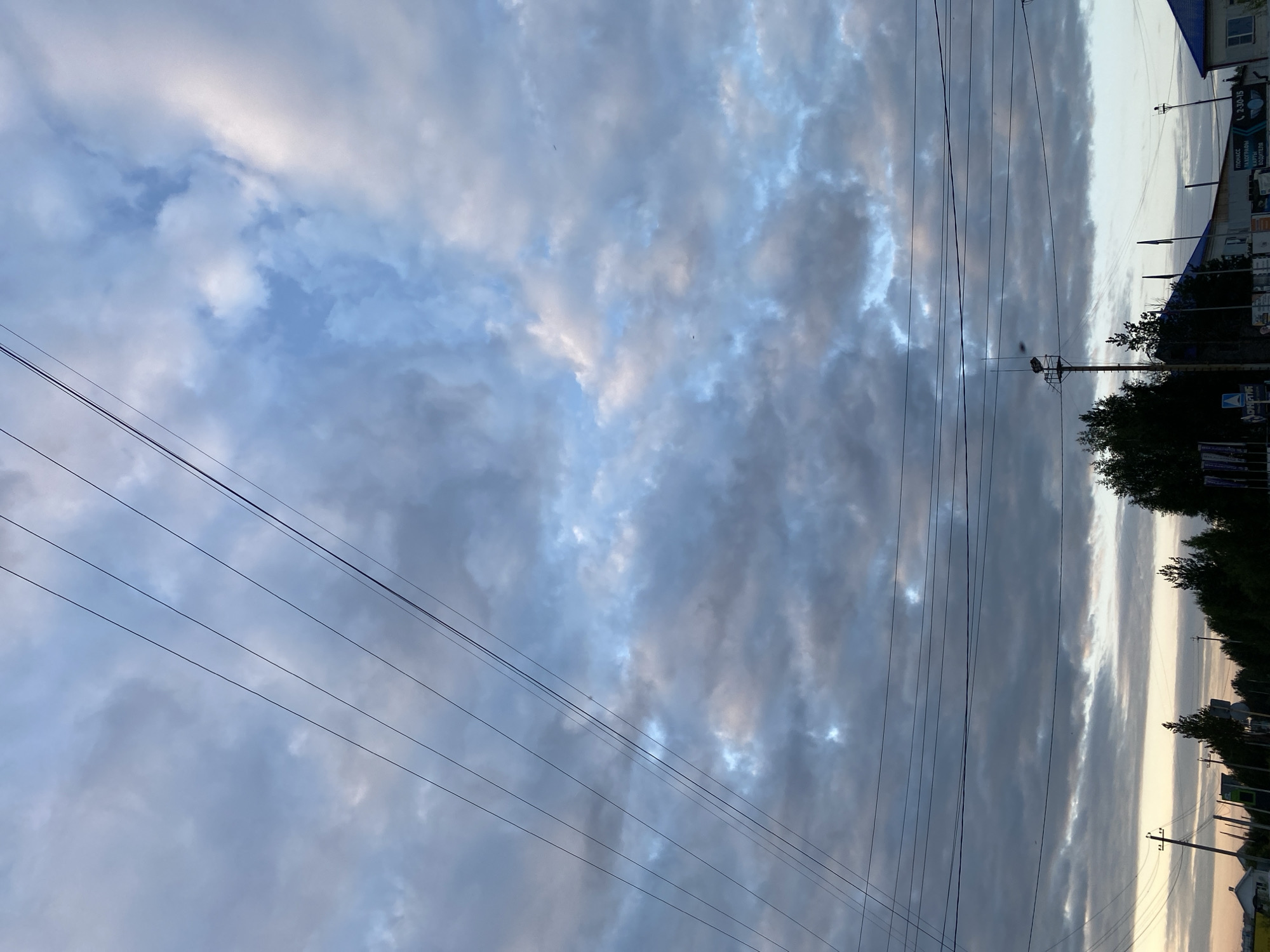 This image depicts a cloudy sky with power lines crisscrossing the frame, seen from a street-level perspective. In the distance, there are trees, buildings, and utility poles, suggesting an urban or semi-urban environment. The lighting indicates it is either dawn or dusk. The scene is consistent with the appearance of a typical street in Nyagan, Russia.