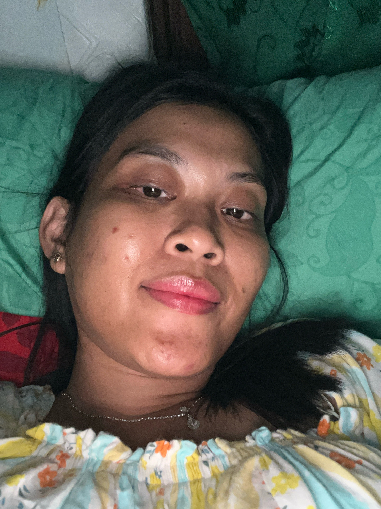 A close-up selfie of a woman lying on a green, patterned bedspread. She has dark hair, brown eyes, and is wearing a floral-patterned top and a delicate silver necklace. The lighting suggests an indoor setting, possibly at night. The background is a blurred green fabric. The image is focused on the woman's face and upper chest.