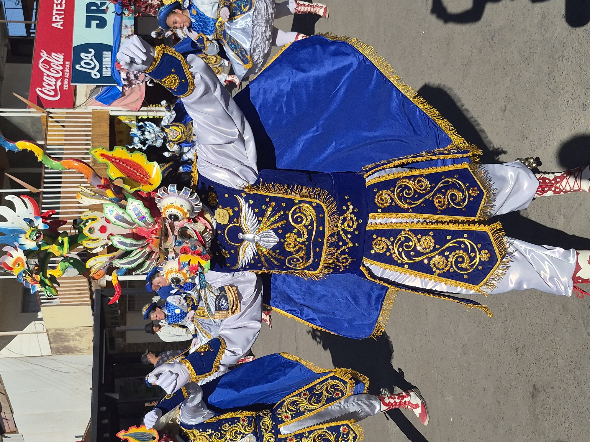 In Pozo Almonte, Chile, a person in a vibrant blue and gold costume with a large, colorful mask resembling a dragon or mythical creature is dancing in the street. The costume features intricate embroidery and dangling gold fringe. The dancer is wearing white pants and red and white boots. In the background, other individuals in similar ceremonial attire are visible, along with buildings and a Coca-Cola advertisement. The ground appears to be asphalt, suggesting an outdoor urban setting. The sunlight casts shadows on the ground, indicating it is daytime.