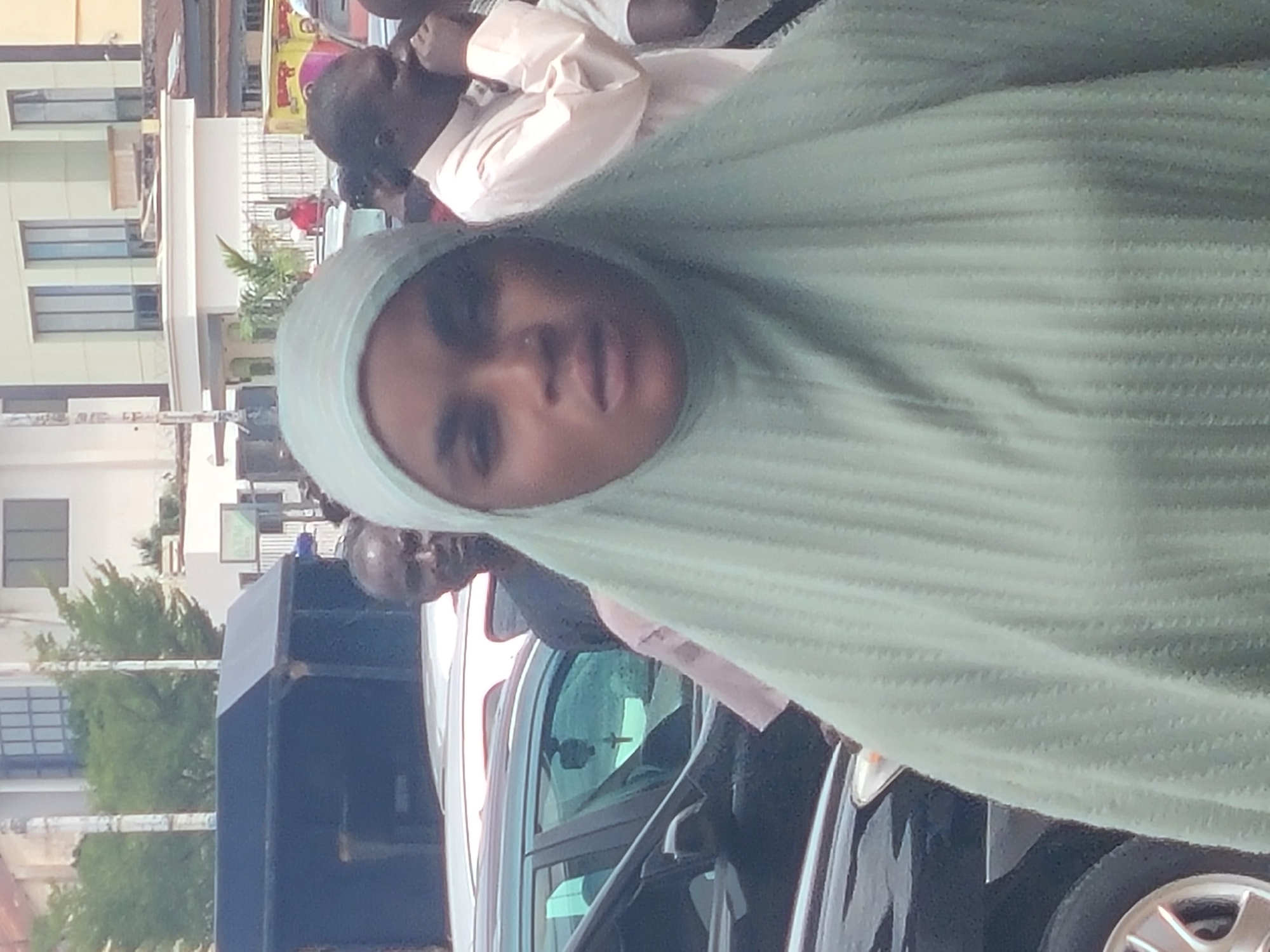 An outdoor, daytime, slightly overexposed image captures a street scene in the city of Unknown, Unknown. The primary subject is a woman of unknown age and ethnicity, occupying the foreground and center, wearing a light-colored (mint green or pale blue) pleated hijab that covers her head and shoulders. She has a nose piercing and her mouth is slightly agape. Behind her, a dark-colored sedan is visible, with a small cross hanging from its rearview mirror. To the main subject's right (viewer's left), the head of a man, also of unknown age and ethnicity, is partially visible. To the main subject's left (viewer's right), another man in a light-colored long-sleeved shirt (possibly peach or pink) is seen with his hand near his face, appearing to be speaking or reacting. In the mid-ground and background, multiple other individuals are present, including a man in a red shirt behind a metal fence. The environment includes several other vehicles (white, red, dark-colored cars), light-colored multi-story buildings with large windows, some green trees, and an ATM sign partially visible on the right. A tall, light-colored pole is present on the left side of the frame. The scene suggests an urban or busy commercial area.