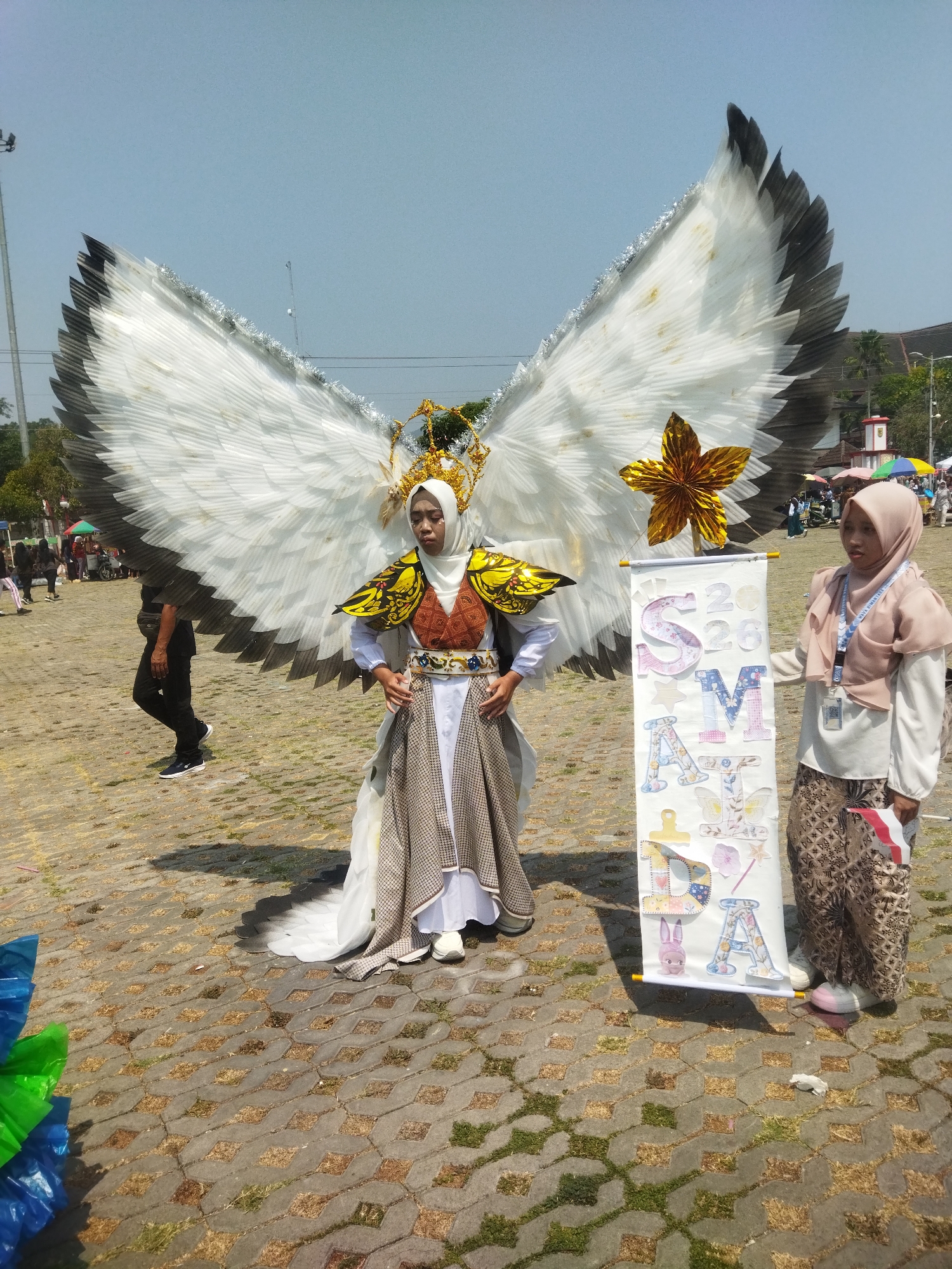 Two young women are posing for a photo at an outdoor event in Wonogiri, Indonesia. The woman on the left is wearing an elaborate costume featuring large white wings with black and silver tips, a golden crown, and a patterned dress. The woman on the right is holding a banner with the letters "S.M.A." and "2026" decorated with colorful designs. The ground is paved with interlocking bricks, and in the background, there are other people, tents, and trees, suggesting a public gathering or festival. The sky is clear and blue.