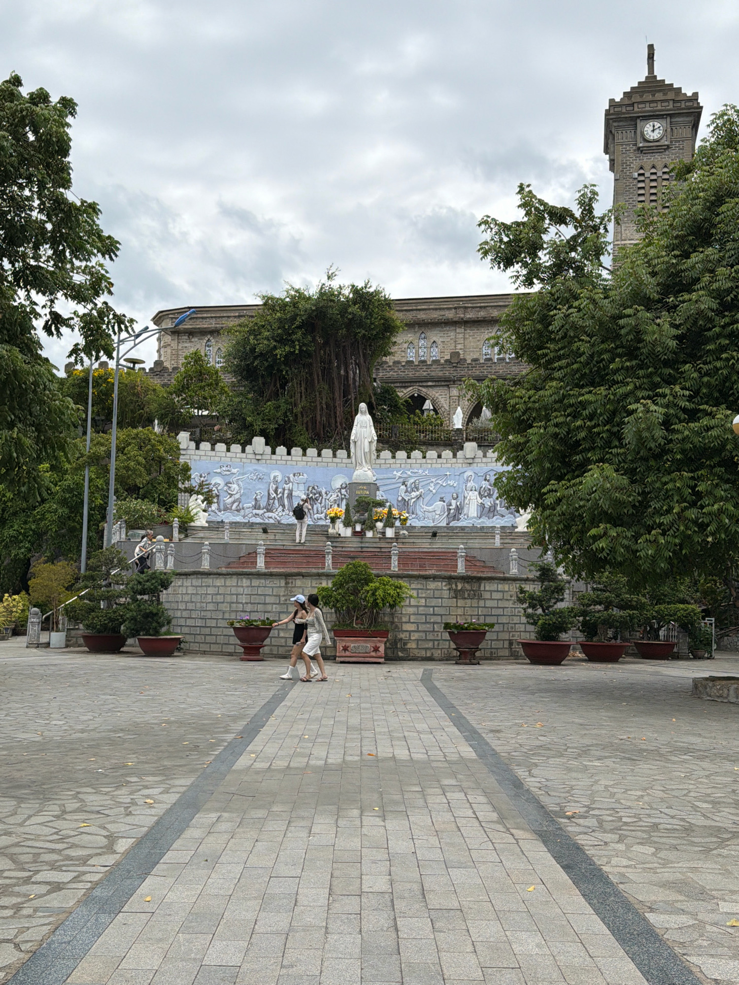 This is a photograph of the Nha Trang Cathedral, a prominent landmark in Nha Trang, Vietnam. The cathedral, constructed in a French Gothic style, features a tall clock tower and an ornate facade. In the foreground, a paved walkway leads towards a series of steps that ascend to a statue of the Virgin Mary. A mural depicting biblical scenes adorns the wall behind the statue. Two young women are walking on the paved area in the foreground. The scene is set against a cloudy sky and surrounded by lush greenery.