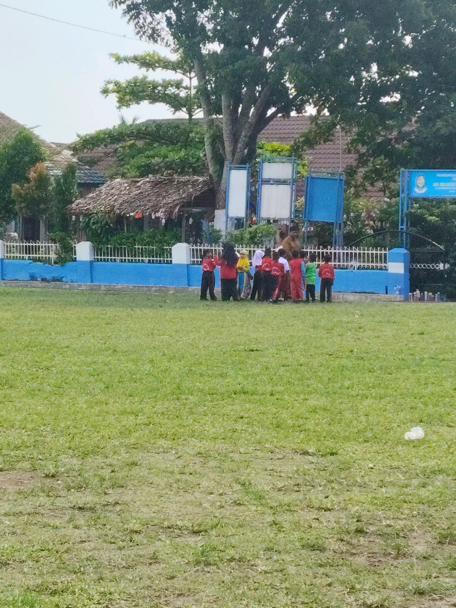 In Sunggal, Indonesia, a group of approximately ten children, some wearing red uniforms, are gathered on a grassy field near a blue and white fence. Behind the fence, a building with a thatched roof is visible, along with trees and two blue signboards. One signboard, on the right side of the image, displays a circular logo with an image and text. The field appears to be a sports or recreational area, with the grass in varying shades of green and some bare patches. The sky is overcast.