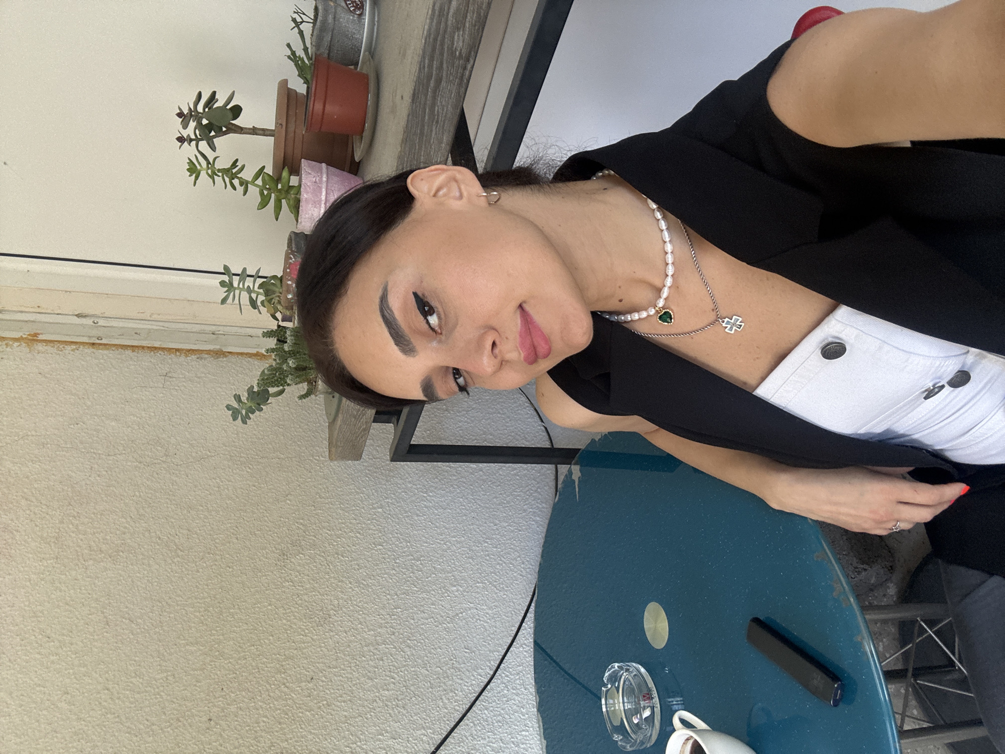 A woman is taking a selfie outdoors at a cafe table. She is wearing a black vest over a white top, and a pearl necklace with a green pendant and a silver cross pendant. A white cup and an ashtray are on the blue table. In the background, there are several potted plants on a shelf. The woman's right hand rests on the table, and her nails are painted red. She is looking at the camera with a slight smile. The wall behind her is textured and cream-colored. The overall setting suggests a casual outdoor seating area, possibly in Tbilisi, Georgia, given the style of architecture and the presence of potted succulents commonly seen in the region.