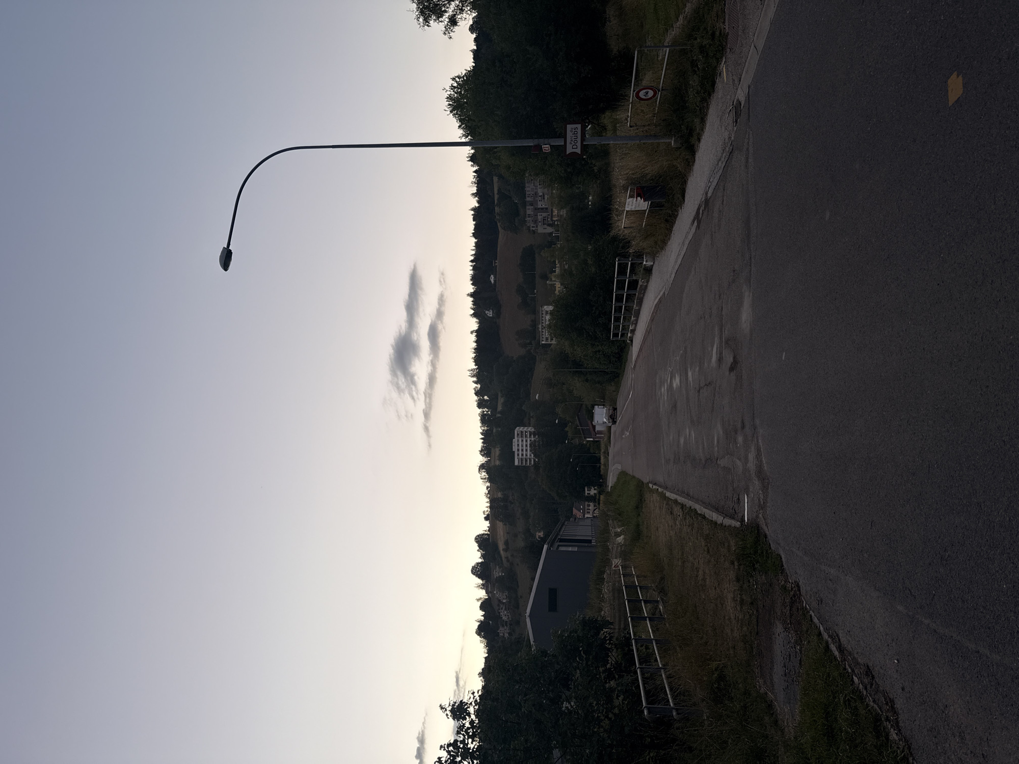 A country road curves towards a hill, lined by a metal fence on the left and a street lamp on the right. The sky is a pale gradient of blue and gray. Buildings, some multi-story, are nestled into the wooded hillside. A dark-colored, rectangular industrial building is prominent on the left. Further down the road, a white recreational vehicle is parked. A signpost with a white sign that reads "Parc du Doubs" and a red circular sign with a bicycle symbol are visible near the street lamp. The road appears to be slightly wet.