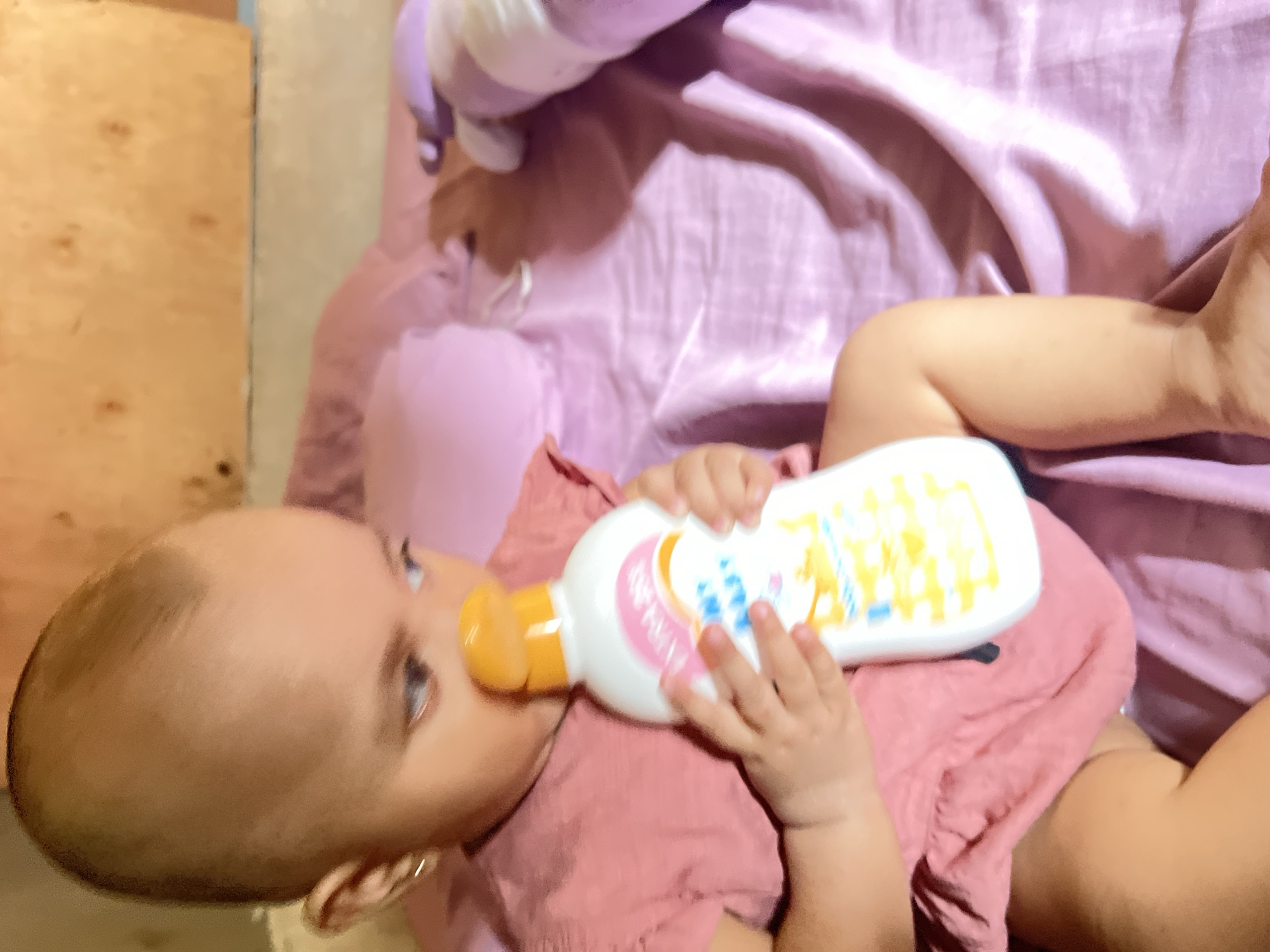 A baby is holding a bottle of baby lotion. The baby is wearing a pink outfit and has a gold earring in her left ear. The baby is looking down at the bottle. The background is a pink blanket. The image appears to be taken indoors, possibly in a bedroom. The lighting is soft and warm. The location is not specified in the image.
