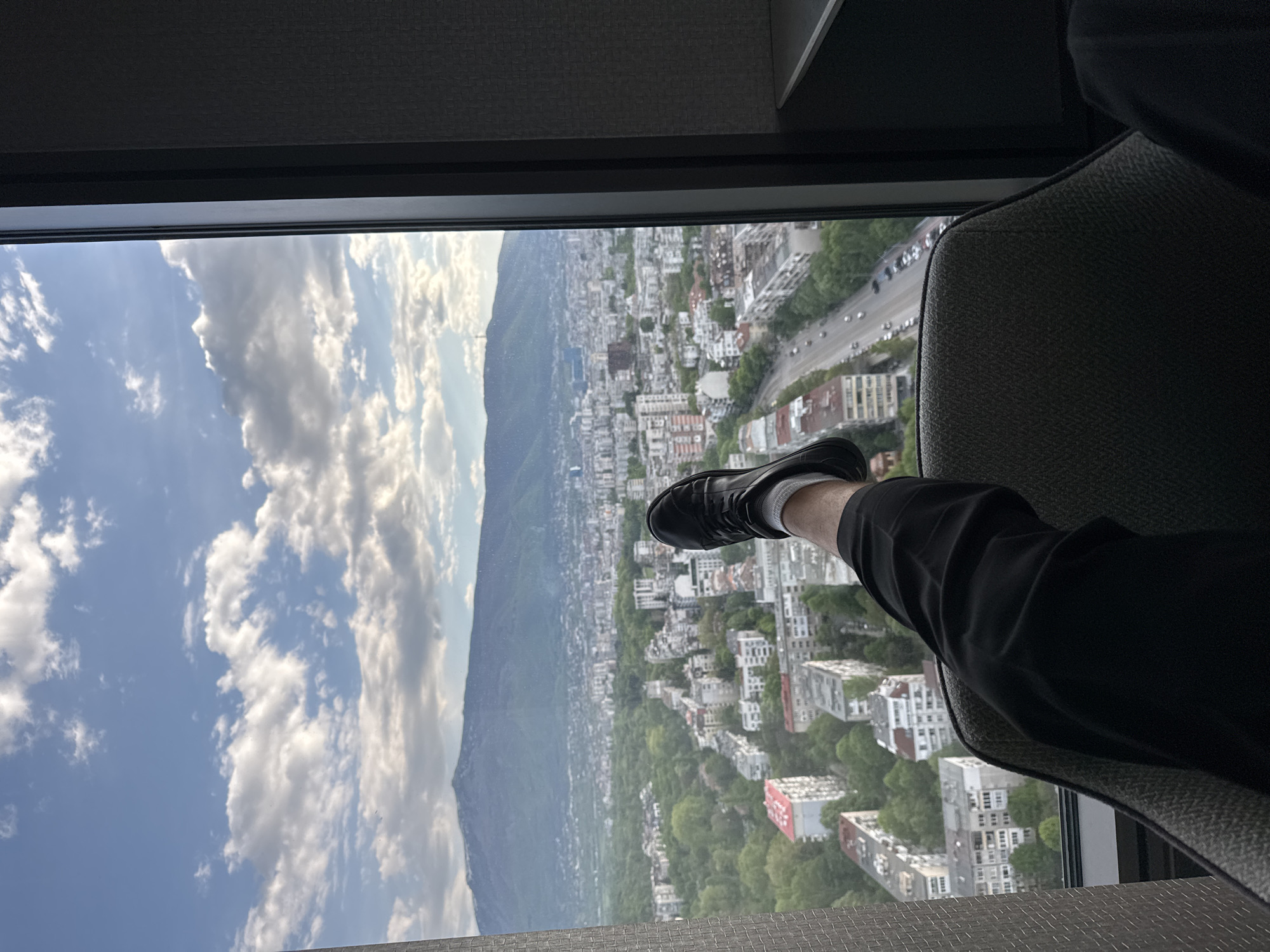 A view through a window in Sofia, Bulgaria, shows a cityscape extending toward a mountain. The sky is partially covered with clouds. A person's leg and foot, wearing a black shoe and white sock, are extended towards the window. The person is sitting in a gray upholstered chair. Buildings and a roadway are visible below. The orientation is rotated 90 degrees counterclockwise.