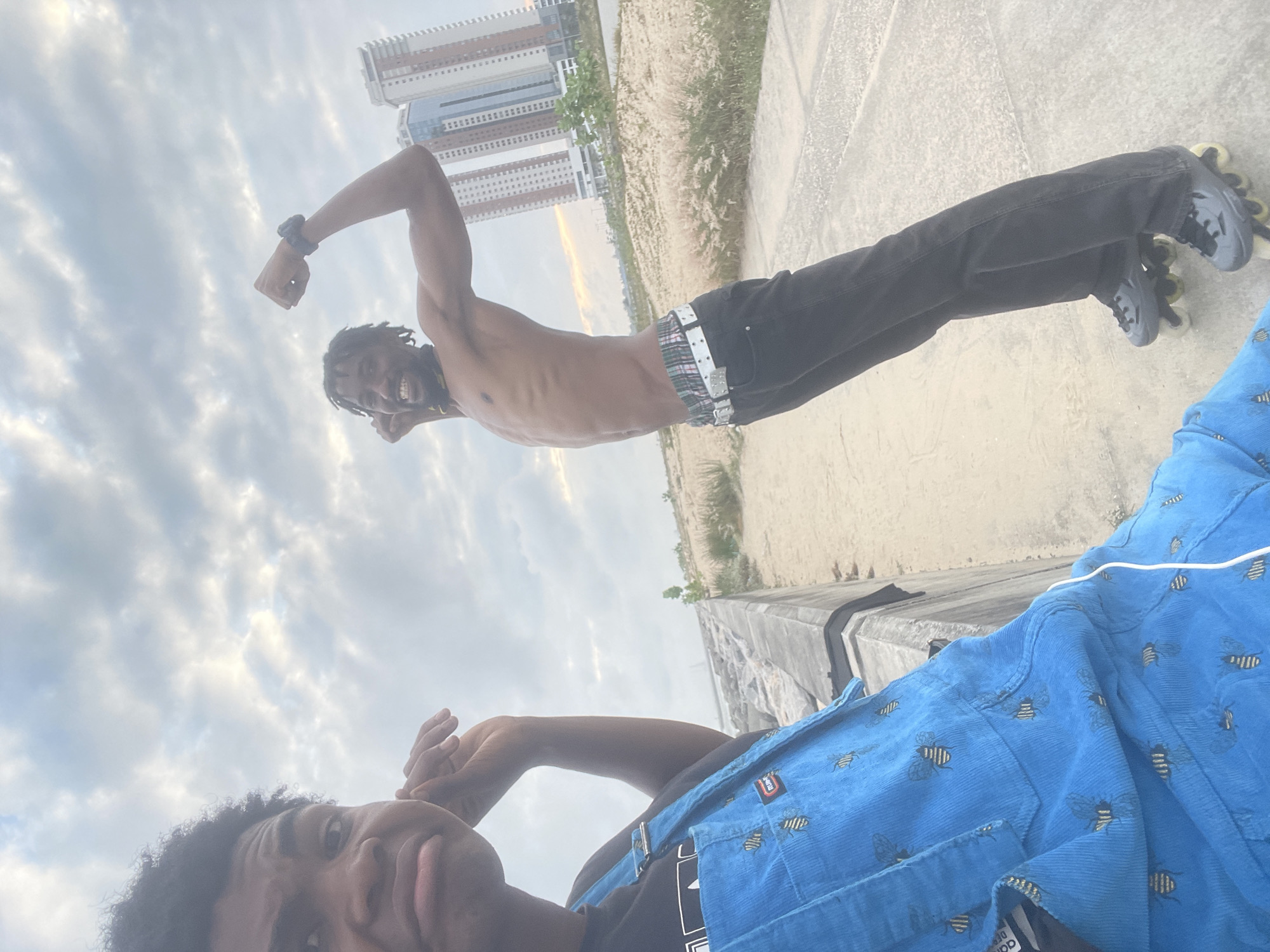 Two individuals are captured in a selfie-style photograph outdoors, likely in the city of Inupa Kekere, Nigeria, given the context of the provided crops. The foreground features a person on the left wearing blue overalls adorned with a bee pattern and a black t-shirt underneath. This individual has dark, curly hair and is looking directly at the camera with a slightly contorted facial expression, with their left arm raised to their ear and temple. The second individual, positioned to the right and slightly behind, is shirtless, revealing a lean physique. They are wearing black jeans with a colorful plaid waistband visible, and rollerblades on their feet. This person is flexing their right bicep and appears to be smiling and looking upwards and to the right.
The background suggests an urban coastal environment. A paved walkway or promenade runs along a sandy area with sparse, dry vegetation. Beyond this, a body of water is visible, with a faint silhouette of a boat on the horizon. Towering modern apartment buildings rise in the distance, indicating an developed urban area. The sky is overcast with dramatic clouds. The lighting suggests it is either early morning or late afternoon. The image appears to have been taken at ground level.