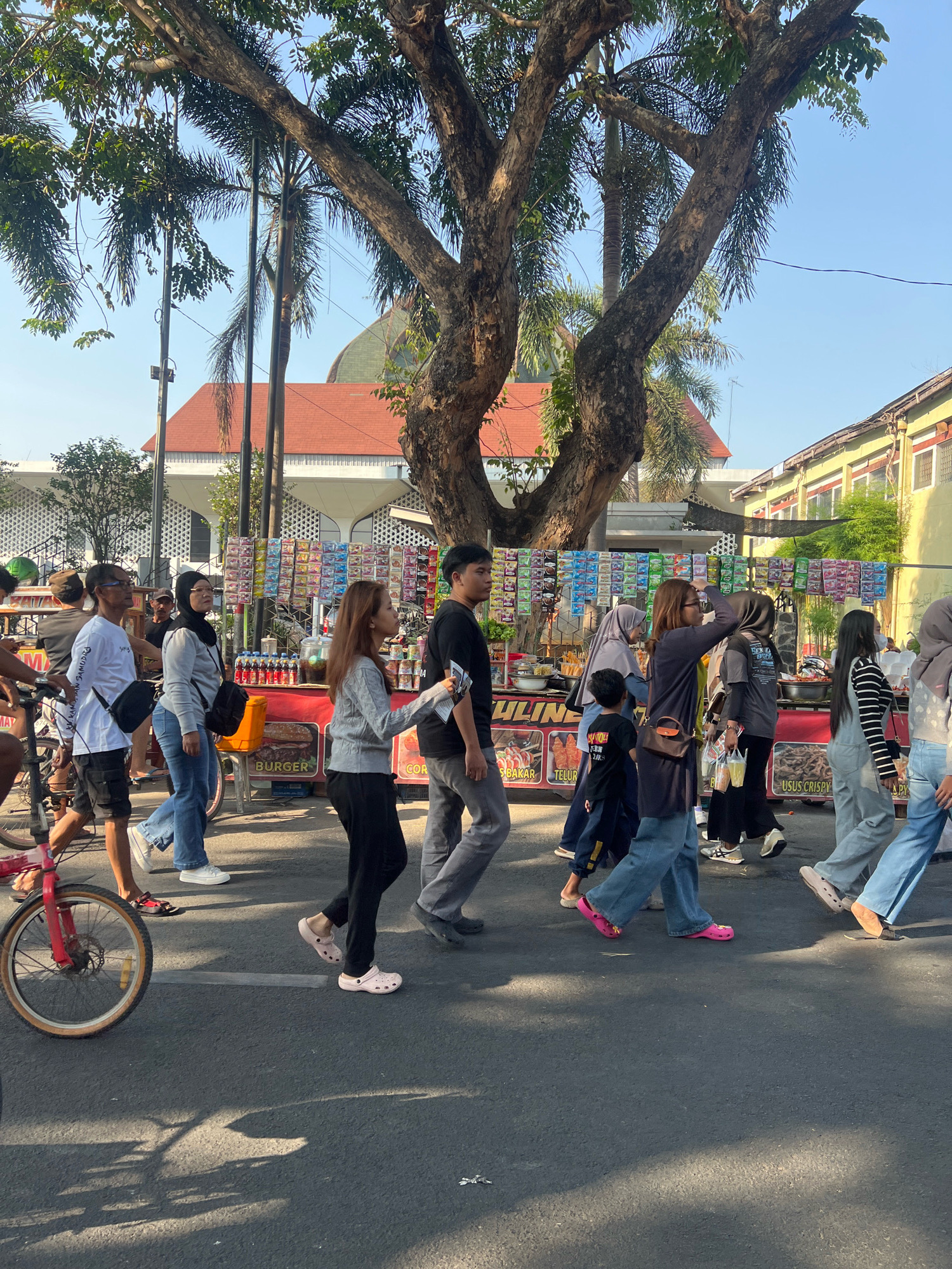 The image depicts a street scene in Kecamatan Pati, Indonesia, on a clear, sunny day. A group of people are walking along the asphalt road, which appears to be a public thoroughfare, possibly a market or festival area. In the background, there are food stalls set up, displaying various items for sale, including what appear to be snacks and drinks. One stall has "BURGER" prominently displayed. Behind the stalls, a building with a red-tiled roof and a green dome is visible, suggesting a cultural or religious site. Large trees with substantial trunks and foliage provide shade over parts of the scene. The people in the foreground are dressed casually in various clothing styles, including jeans, t-shirts, and headscarves, indicating a diverse group of individuals. Some are carrying bags, and one person is pushing a bicycle with a red frame. The overall atmosphere suggests a lively outdoor gathering or a busy shopping street.