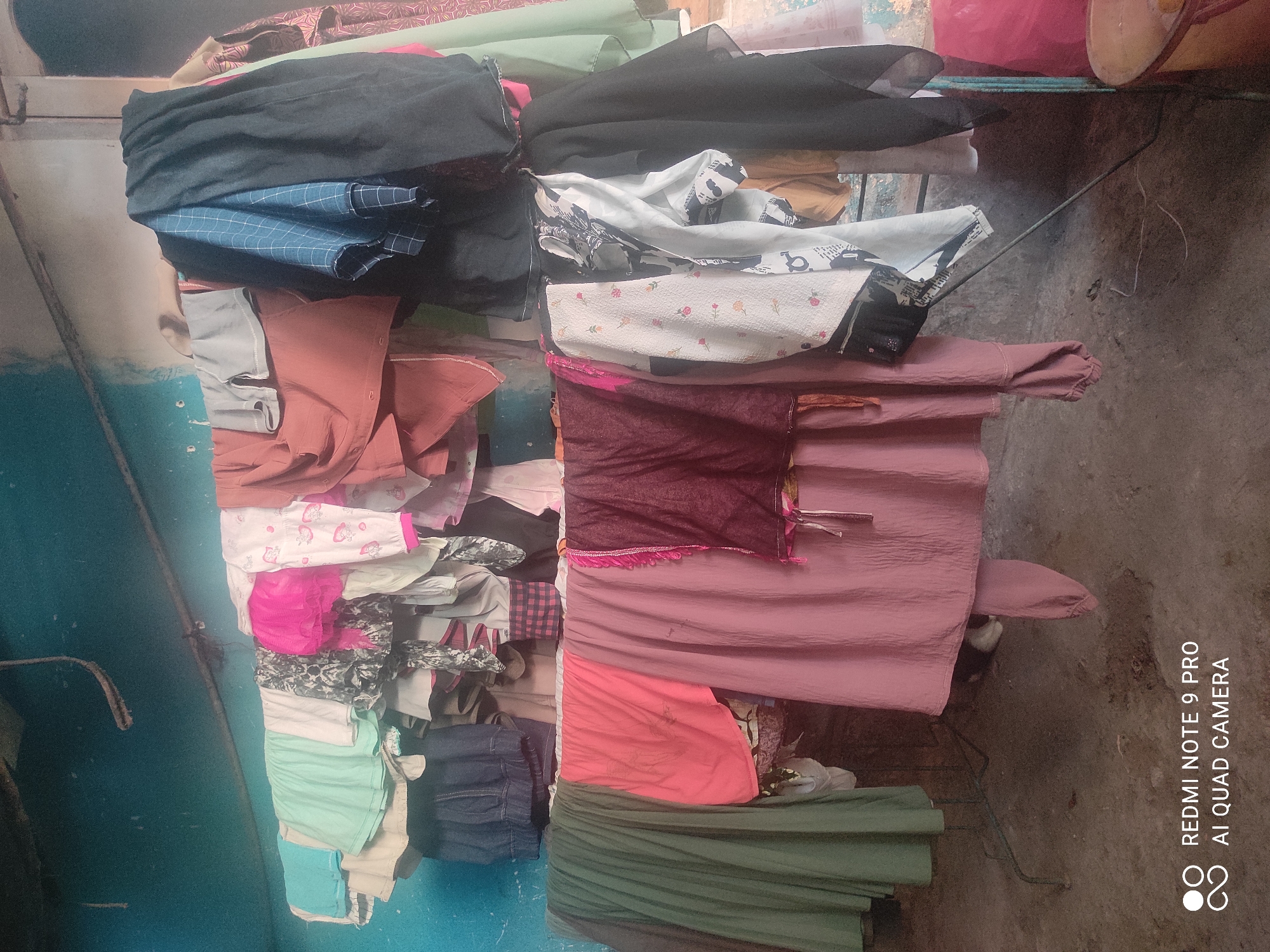 In Sakra, Indonesia, a clothesline is strung across a room with blue walls. Various articles of clothing, including skirts, shirts, and pants, are hung to dry. The clothing ranges in color from light blue and green to pink, red, black, and white with patterns. Some of the items are folded, while others hang loosely. A rack and a metal pole are visible supporting the clothesline. The floor is made of concrete. In the lower right corner, a partially visible yellow bucket and a pink plastic object are present. The overall impression is one of domestic laundry being air-dried.