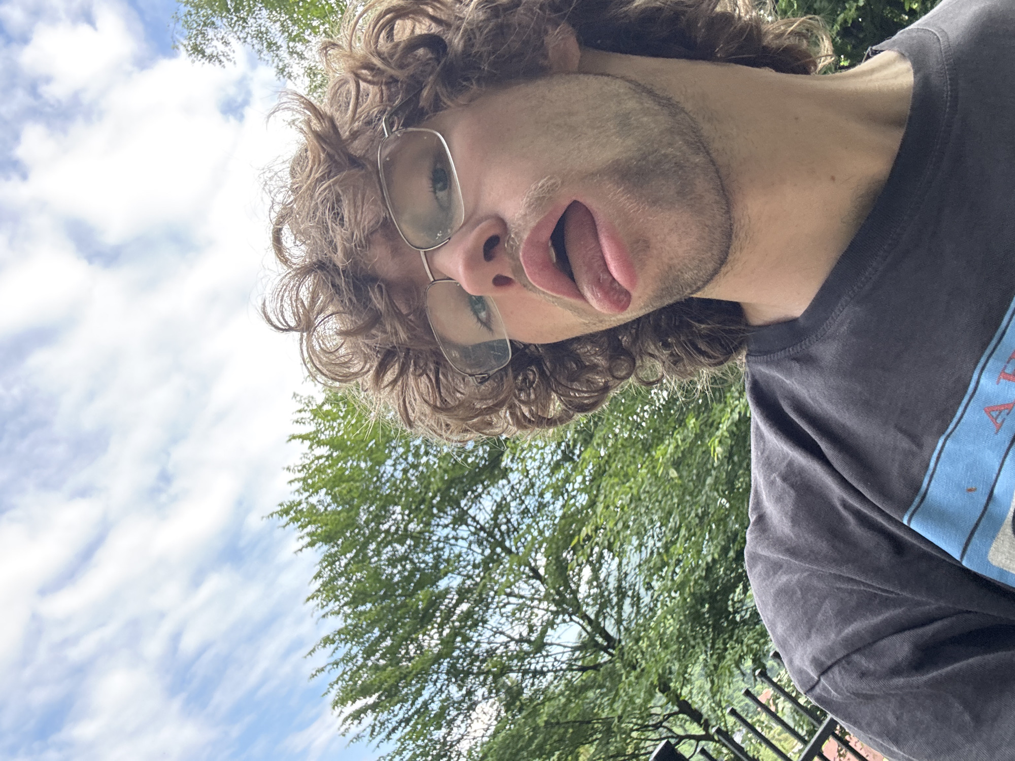 An extreme close-up outdoor selfie shows a young adult male with curly brown hair and glasses sticking out his tongue at the camera. He is wearing a dark gray t-shirt with a blue and black stripe design across the chest. In the background, out of focus, are green trees and a blue sky with white clouds. The lower left corner of the frame shows a black metal fence. The image appears to have been taken from a low angle looking up at the subject.