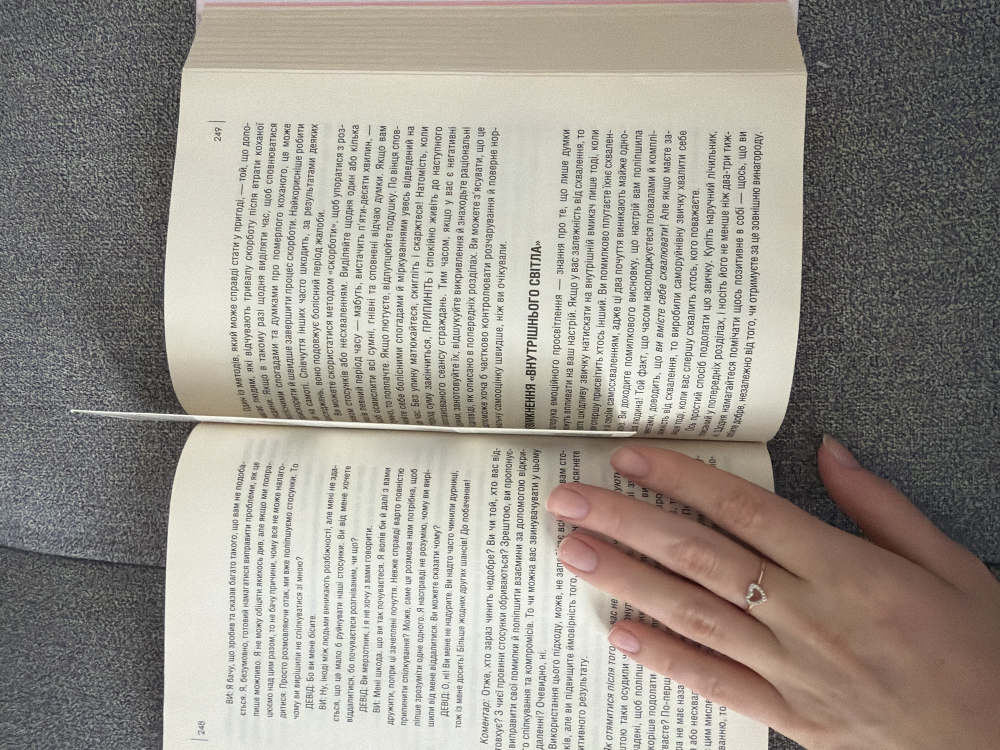 The image shows an open book lying on a gray textured surface, possibly a table or floor. A person's left hand, adorned with a delicate ring shaped like a heart with small stones, rests on the right-hand page of the book. The hand is positioned in the lower right quadrant of the image, partially obscuring the text. The book appears to be a paperback, with text printed in Ukrainian. The pages are slightly bent, suggesting it has been recently read or handled. The lighting suggests an indoor setting, and there are no visible indications of the city of Kyiv, Ukraine, or any specific location.