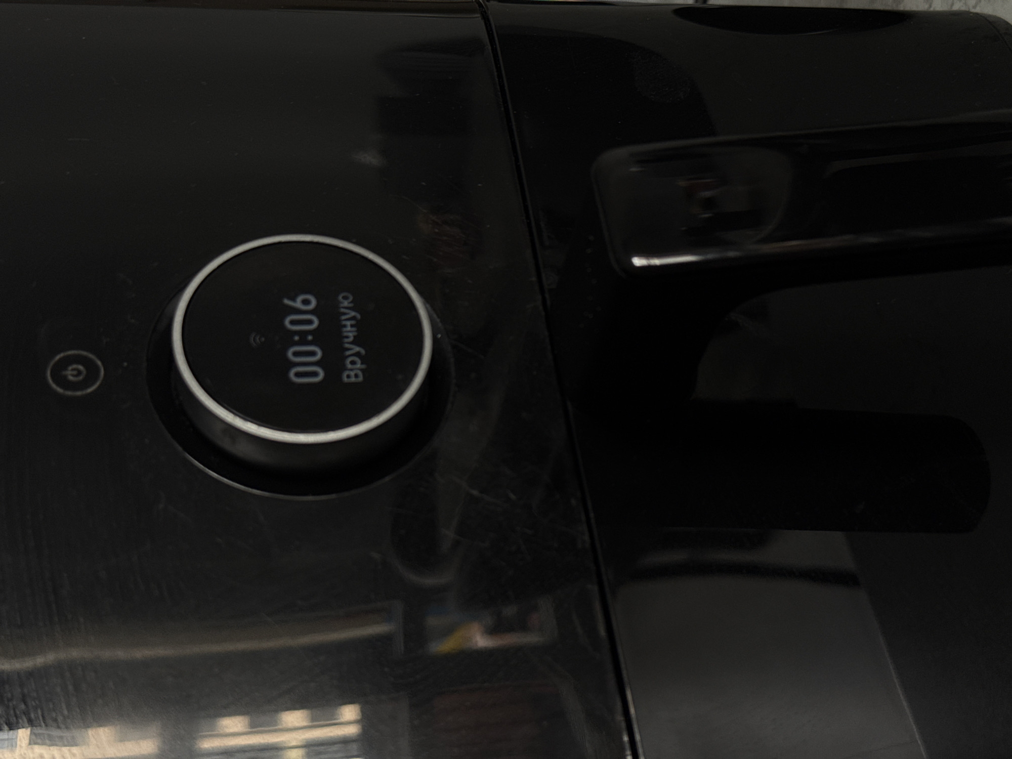 The image shows a close-up of a black appliance, likely an air fryer or similar kitchen device. The top of the appliance features a circular digital display showing "00:06" and text below it that reads "Вручную" in Cyrillic script, which translates to "Manually". Above the display is a power button icon. The appliance has a glossy finish with visible scratches and smudges. A dark, reflective handle is visible in the foreground on the right side of the image. The background is dark and out of focus, with some vertical lines that might be reflections from window blinds or similar objects. There are no people or animals present in the image. The location is not determinable from the image alone; it is not possible to determine if this is in Kraskovo, Russia.