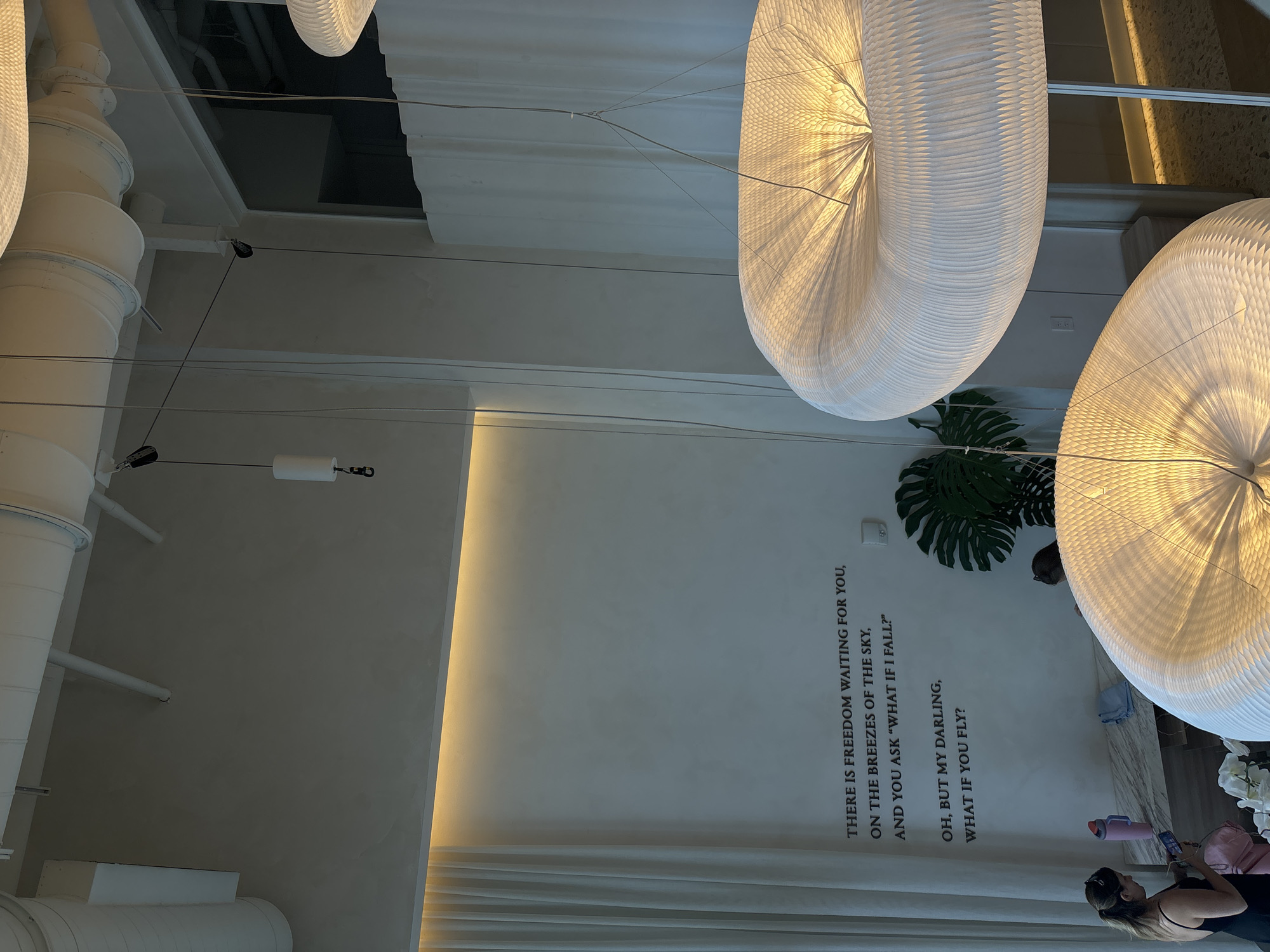 The image appears to be an indoor shot taken from a low angle, looking upwards in a building. The main visual elements are large, illuminated, decorative hanging lamps that resemble inflated paper lanterns. A white ceiling and wall dominate the background, with long white pipes running across the ceiling.
On the lower right side of the wall, there's a printed phrase "THERE IS FREEDOM WAITING FOR YOU, ON THE BREEZES OF THE SKY, AND YOU ASK 'WHAT IF I FALL?' OH, BUT MY DARLING, WHAT IF YOU FLY?". A woman can be seen at the very bottom right corner of the image, possibly using a mobile phone.
The building's location is suggested to be in Miami, United States of America.