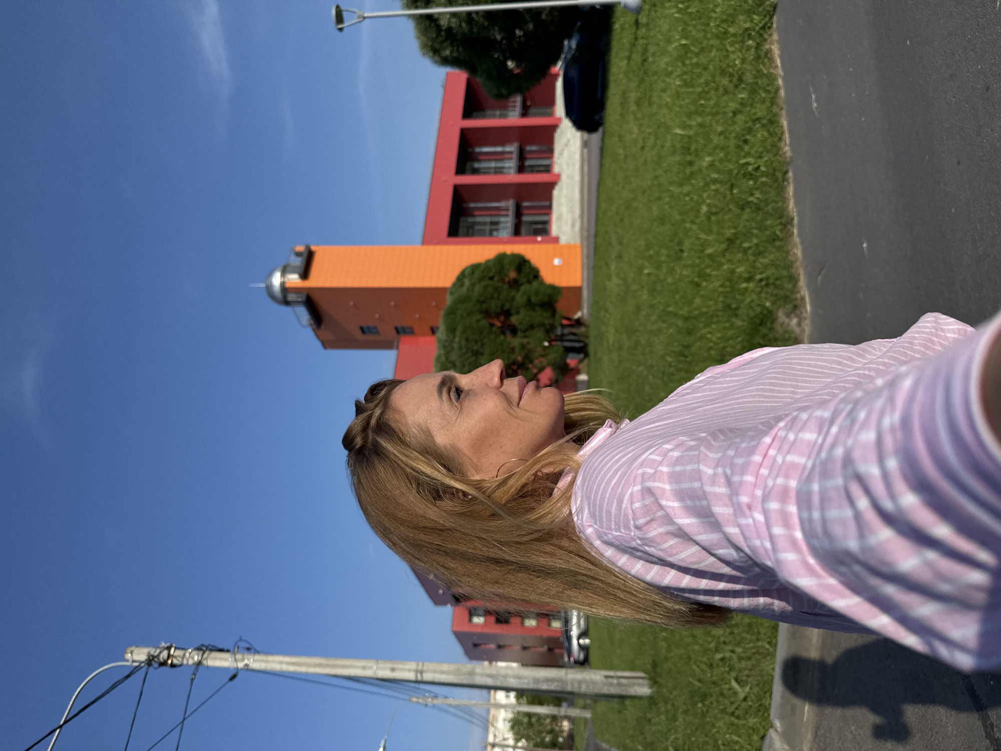 A woman is photographed from the chest up, in profile, facing right. She has blonde hair with auburn highlights, pulled back into a bun on top of her head. She is wearing a pink and white striped collared shirt. The background shows a bright blue sky, green grass, a modern orange and red building complex with a tall orange tower, and a utility pole with wires on the left. A black car is parked on the right side of the image. This image was taken in the city of Kirishi, Russia.