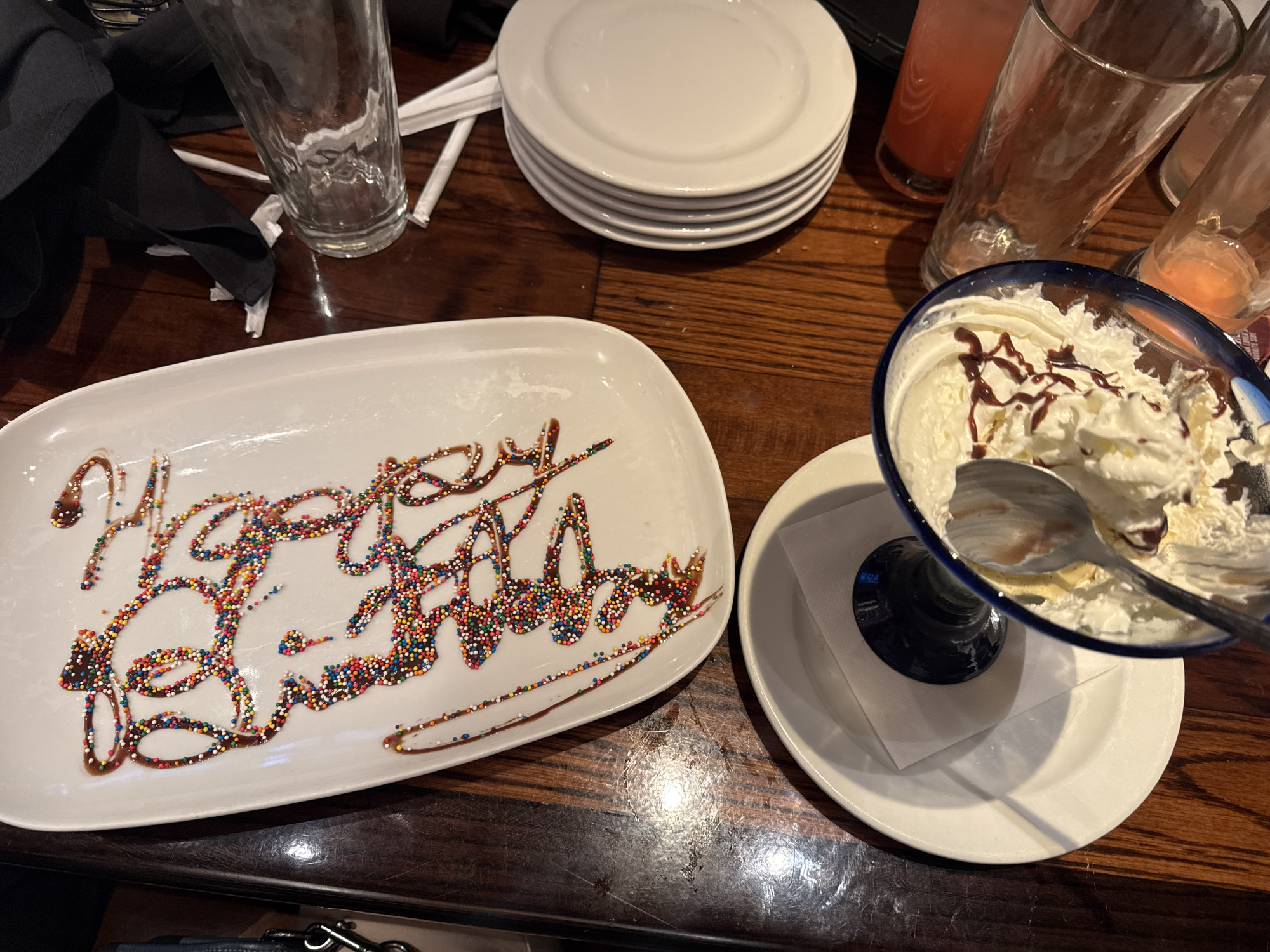 The image displays a tabletop scene, likely in a restaurant in Schaumburg, United States, captured after a birthday celebration. The central focus is a white, rectangular ceramic platter on which the words "Happy Birthday" are artfully written in chocolate syrup, heavily coated with vibrant, multi-colored sprinkles.
To the right of the birthday plate, a blue-rimmed glass dessert bowl contains the remnants of a sweet treat, primarily whipped cream with streaks of chocolate syrup, and a spoon resting inside, indicating it has been mostly eaten. This dessert bowl is placed on a small white saucer with a folded white napkin underneath.
The table is made of dark wood, showing reflections from the lighting. In the background, a dark cloth napkin is casually placed near an empty, clear drinking glass and some white paper items. A stack of five clean white round plates is neatly arranged further back on the left. On the right side of the background, several other empty or near-empty drinking glasses are visible, some with traces of a light pink or orange beverage. No people are present in the frame, but the arrangement suggests a recently concluded festive gathering.