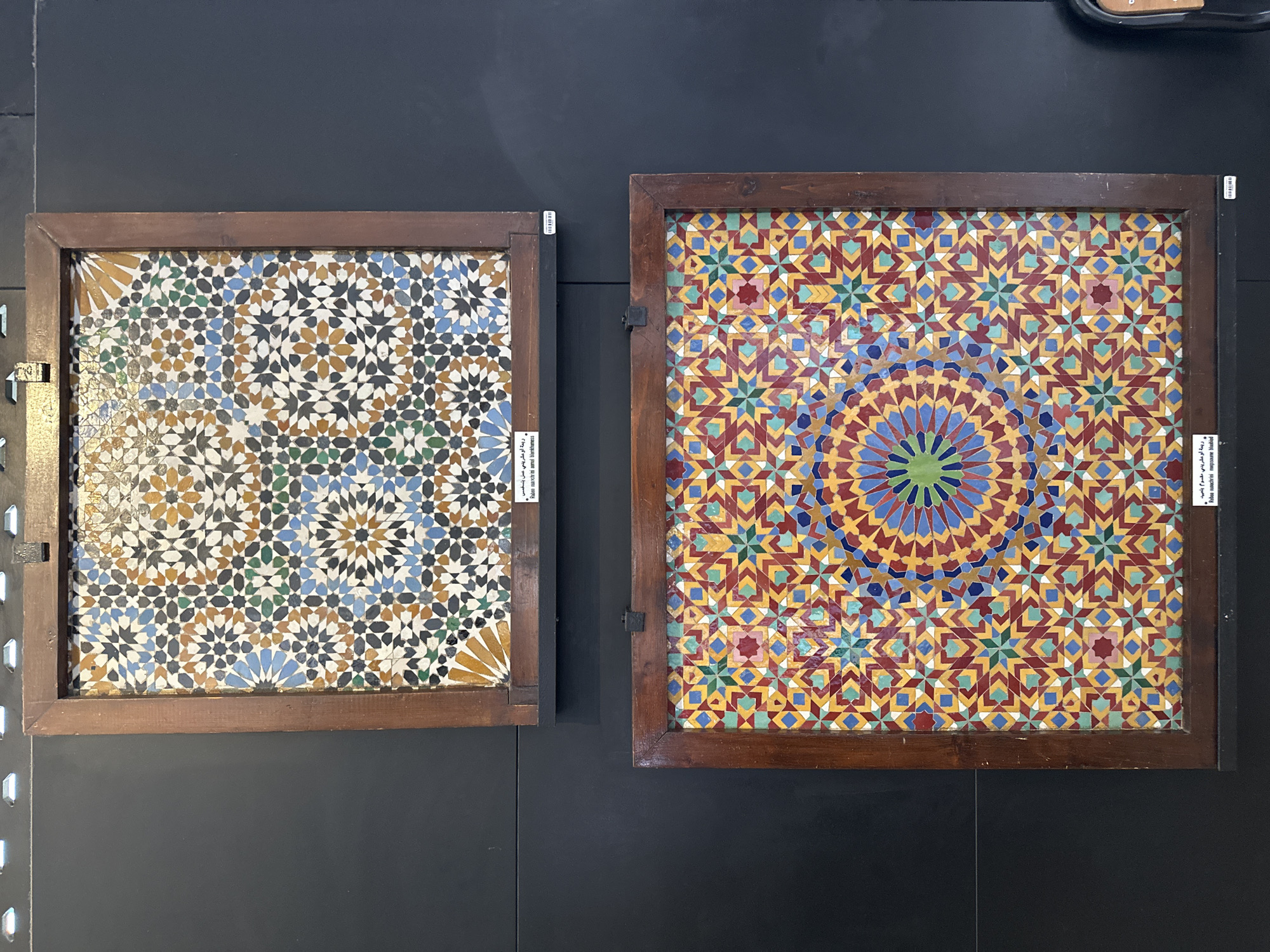 The image displays two framed panels of traditional Moroccan zellige mosaic tiles, presented against a plain dark gray wall, likely in a museum or exhibition setting in Casablanca, Morocco.
The panel on the left features a complex geometric pattern composed of tessellated multi-pointed stars and various polygons. Its color palette is dominated by earthy tones including shades of brown, beige, cream, dark green, and black, with notable accents of light and dark blue.
The panel on the right showcases a more vibrant and elaborate geometric design. It features a prominent central circular motif with green and blue segments, from which a radiating pattern extends across the entire panel. This larger pattern incorporates a rich array of colors, including red, yellow, blue, green, and orange stars and polygons.
Both mosaic panels are encased in simple, dark-stained wooden frames that appear somewhat aged. Each frame has a small white informational label attached to its right side. The label on the left panel displays text in Arabic and Latin script, reading: "ربعة أو عشريني عمل بلمخمس" and "Rabea ouanchrini aamel blamkhamess". The label on the right panel also features text in both scripts: "ربعة أو عشريدي مقسوم بلعبيد" and "Reboa quachrini magsoume blaabied". The overall scene highlights the intricate artistry and mathematical precision inherent in traditional Moroccan zellige craftsmanship. No people are present, and no specific event or activity is occurring beyond the static display of these artworks.
