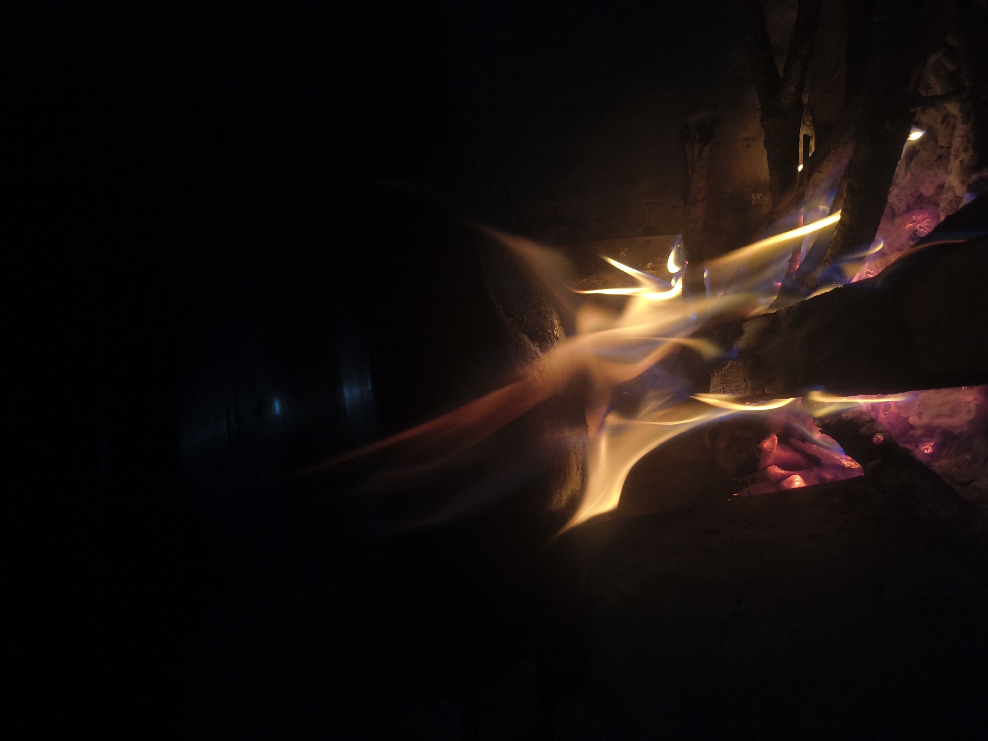 A low-angle, medium shot captures a close-up view of a campfire at night. The flames, predominantly in shades of yellow, orange, and blue, lick upwards from burning logs. The scene is dark, with the fire providing the main source of light. In the background, the darkness is partially broken by a faint, bluish, possibly reflective surface, hinting at the presence of a ceiling or overhang. The environment appears to be an enclosed space, possibly a hearth or a makeshift fire pit, with dark, rough textures visible on the left and right edges, suggesting stone or brick. No people are visible in the frame. The image is a static representation of the dynamic dance of flames. The specific location in Haini, India, is not discernible from the visual information provided.