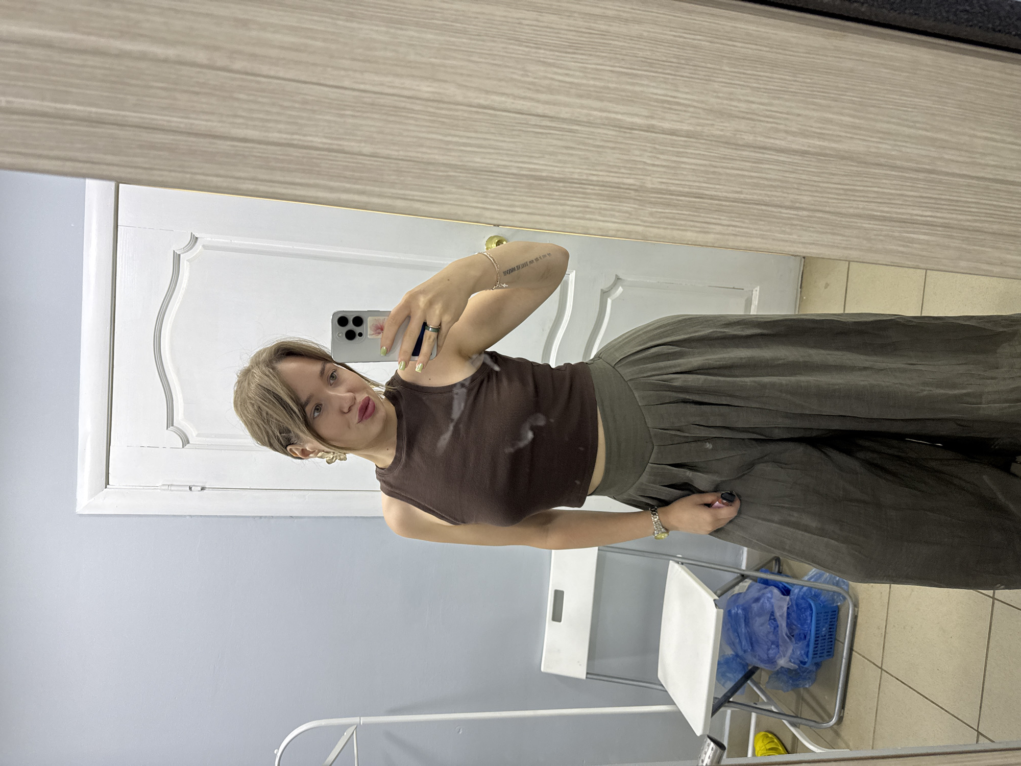 A young Caucasian woman with blonde hair is taking a mirror selfie in what appears to be a room with a tiled floor and a light blue wall. She is wearing a brown ribbed tank top and wide-leg olive green pants. She has large gold earrings and is wearing a watch on her left wrist and a ring on her right index finger. She is holding a smartphone in her right hand and has her left hand on her hip. In the background, a white folding chair is visible, with blue plastic bags underneath it. A white door with decorative molding is behind the chair. The image was taken in Chaikovsky, Russia.