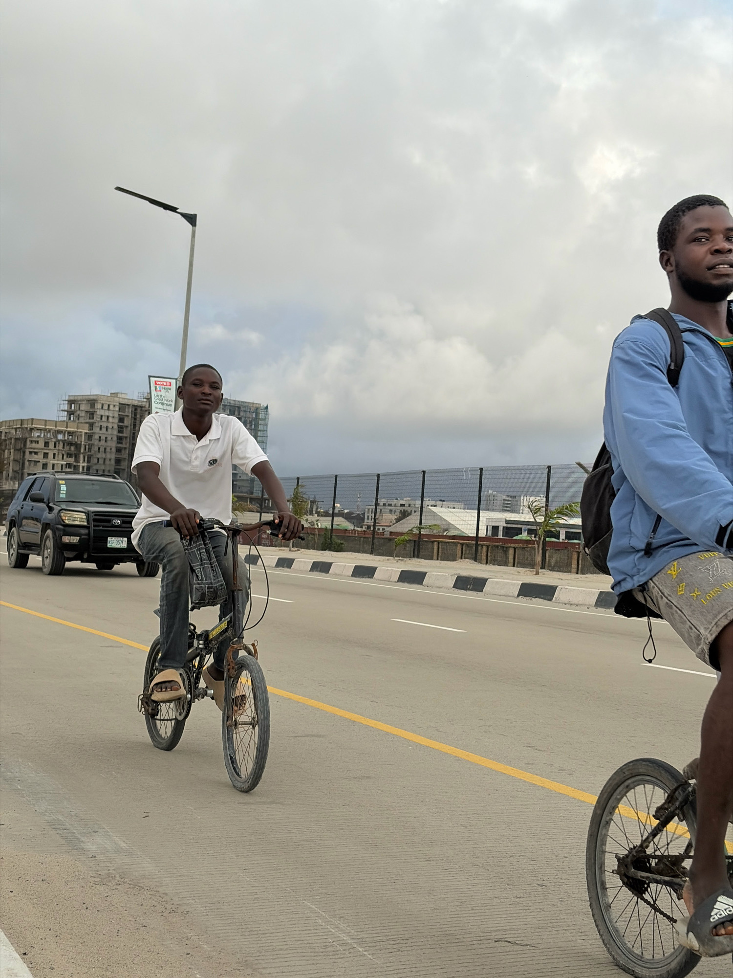 Two young Black men are riding bicycles on a paved road in Alaguntan, Nigeria. The man on the left, in the foreground, wears a white polo shirt and dark jeans, riding a small, black bicycle with a bag hanging from the handlebars. The man on the right, partially visible and further down the road, wears a blue jacket and patterned shorts, also riding a bicycle and carrying a black backpack. They are both facing forward, suggesting they are traveling in the same direction. The road is bordered by a yellow line on the left and a black and white striped curb on the right. In the background, there is a black SUV, unfinished buildings, a lamppost, and a fence. The sky is overcast with clouds.