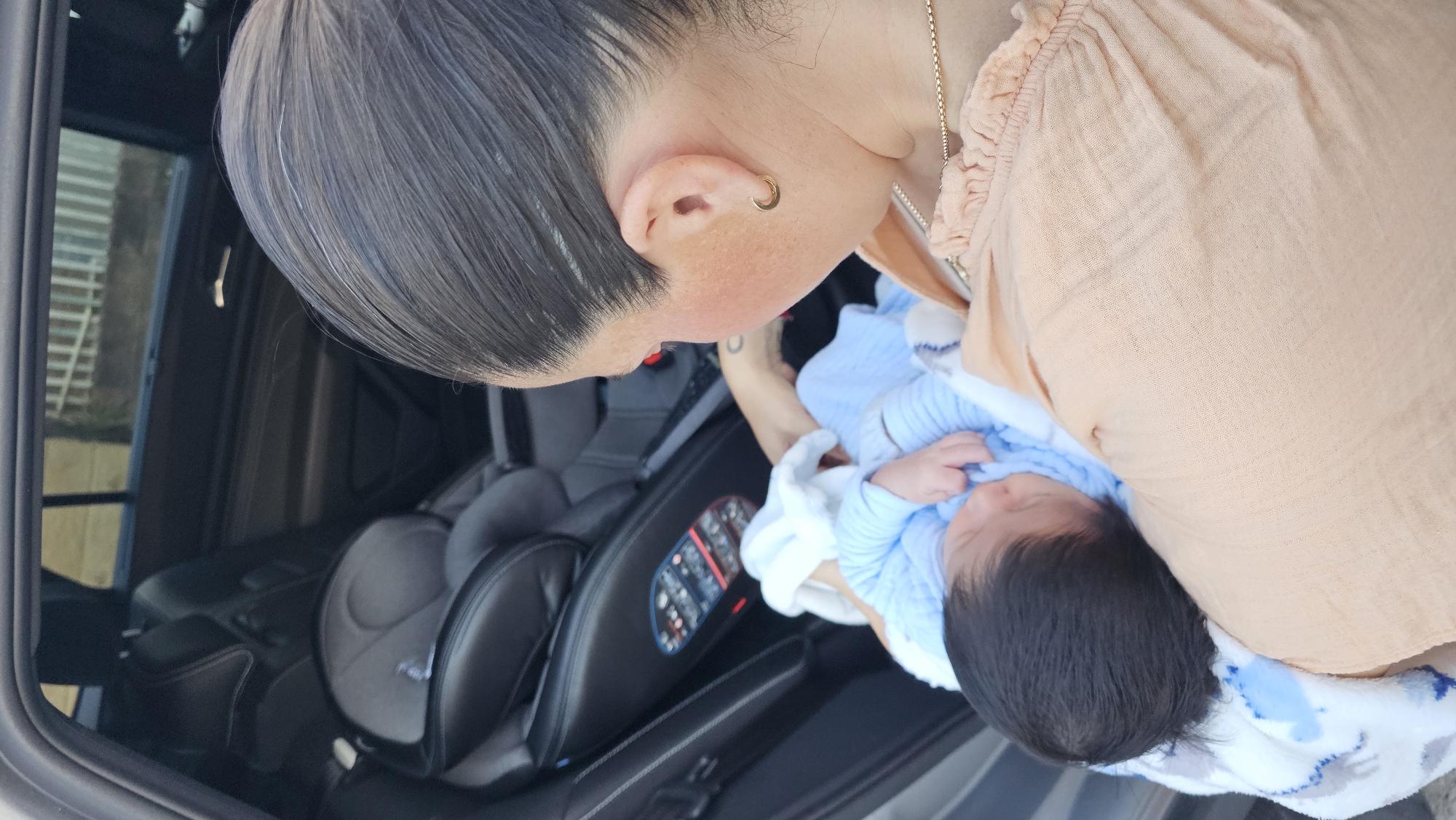 A person wearing a peach-colored blouse is holding a baby wrapped in a blue and white blanket. They are standing beside a car seat inside of a vehicle. The photo was likely taken in San Bernardino Tlaxcalancingo, Mexico.