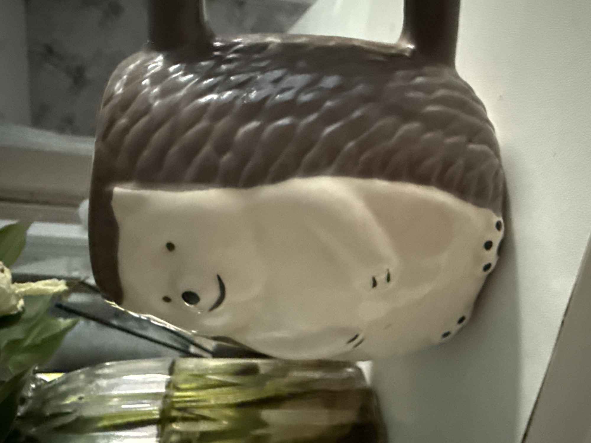 The image shows a close-up of a ceramic mug shaped like a hedgehog. The mug is light brown with a textured pattern representing quills on its back and a cream-colored belly with black dots for feet. It has a handle on the right side and a cute, smiling face with black eyes and a curved mouth. To the left of the mug, a portion of a glass vase filled with greenery and water is visible. The mug and vase are placed on a white surface, possibly a shelf or table, in what appears to be an indoor setting. The background is out of focus, showing muted tones and textures. There is no indication of the city of Октябрьское, Russia, in the image.