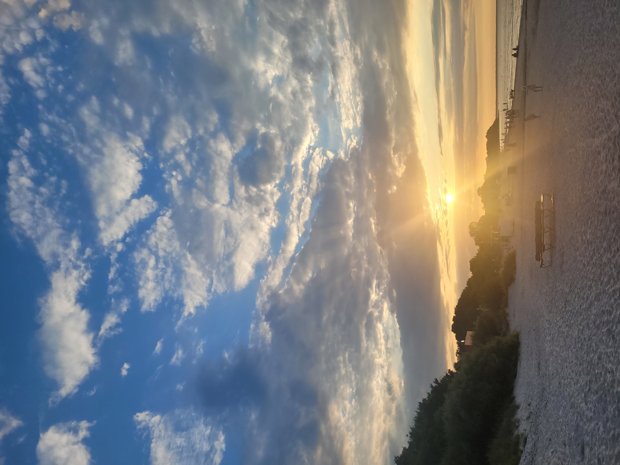 The image captures a beach scene at sunset in Jūrmala, Latvia. The sky is a dramatic display of blue with scattered white and gray clouds, illuminated by the warm, golden glow of the setting sun. Rays of sunlight pierce through the clouds, casting a soft light over the scene. The beach is sandy, with a gentle slope towards the calm sea. On the left side, a dense line of green trees and bushes borders the beach, with some buildings subtly visible behind them. In the foreground, empty park benches made of wood and metal are positioned on the sand. Several people are present on the beach, spread out across the sand and near the water's edge. Some are walking, while others are seated or standing, enjoying the evening ambiance. The overall impression is one of tranquility and natural beauty.