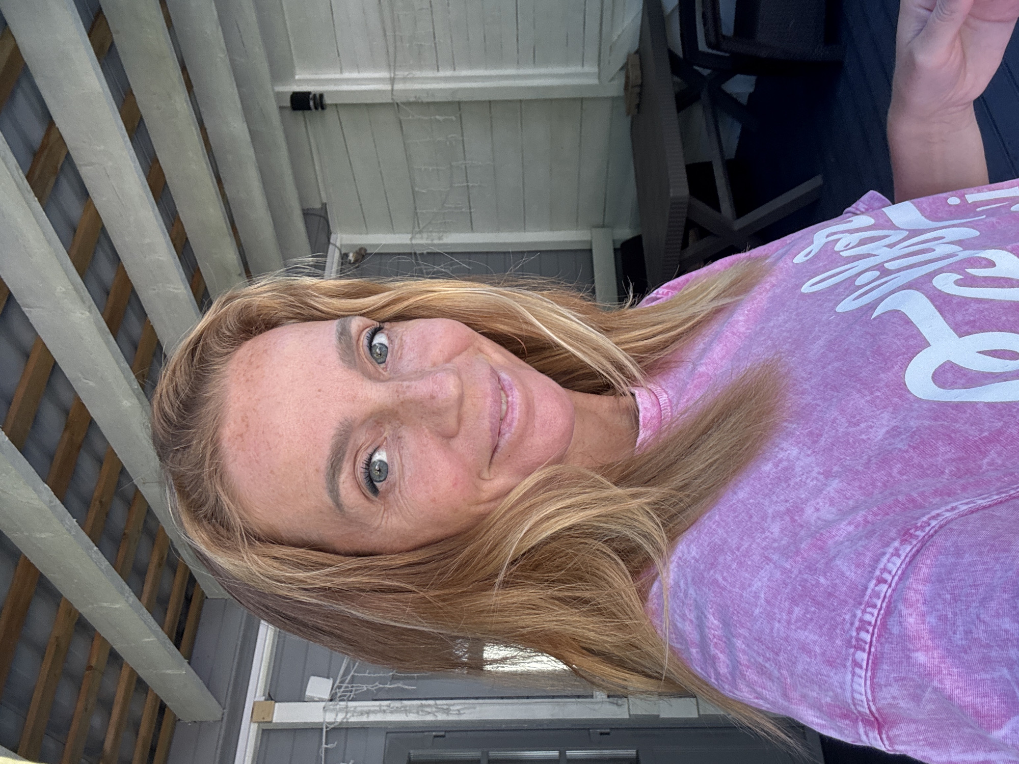 An outdoor, eye-level, medium shot shows a woman with long, wavy blonde hair and freckles on her face, wearing a pink tie-dye t-shirt, taking a selfie. She is smiling with her lips slightly parted. Behind her, a covered porch with exposed wooden beams and white siding is visible. A picnic table and chairs are to the right of the frame. The image appears to have been taken during the daytime in the city of Zelenaya Roshcha, Russia.