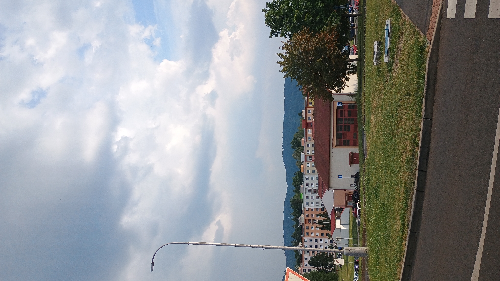 This is an outdoor, ground-level, vertical, full-shot photograph taken on a partly cloudy day in Sokolov, Czechia. In the foreground, a paved road runs horizontally across the bottom of the frame, with a grey curb and a grassy median to its right. The median contains two rectangular, white, solar-powered lights. Behind the median is a patch of green grass. In the middle ground, a single-story building with a red-tiled roof and large red doors, possibly a fire station, is visible. To the left of this building are several smaller white structures. Further back, a long, multi-story residential building with colorful facades (orange, white, and green) spans across the mid-ground, nestled against a backdrop of forested hills. A tall, slender lamppost stands on the left side of the image, extending from the bottom towards the sky. The sky is filled with scattered white and grey clouds.