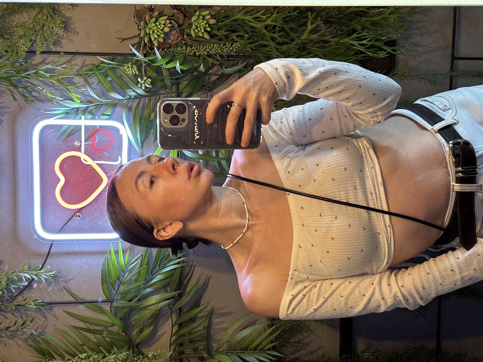 A woman takes a selfie in front of a wall decorated with artificial plants and a neon sign shaped like a social media icon with a heart. She is wearing a patterned white crop top with long sleeves, light-colored denim jeans with a dark leather belt, and a beaded necklace. The city of Votkinsk, Russia, is not visible in the image.