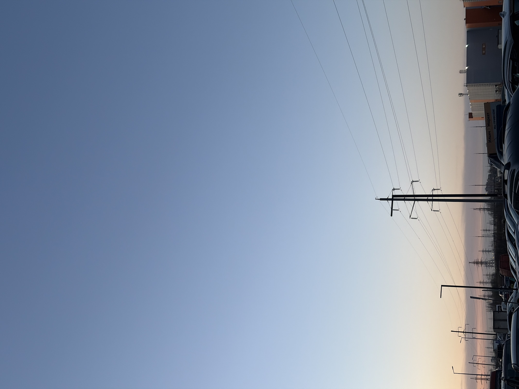 The image shows a view towards the horizon in what appears to be an industrial or commercial area. The sky is a gradient of blue to light orange, suggesting either dawn or dusk. A power transmission tower stands prominently in the middle-right of the frame, with several power lines stretching across the upper portion of the image. To the right of the tower, there are buildings and what appears to be a parking lot filled with cars. Street lights are visible. The location may be Saint Petersburg, Russia, but further landmarks would be required for definitive identification.
