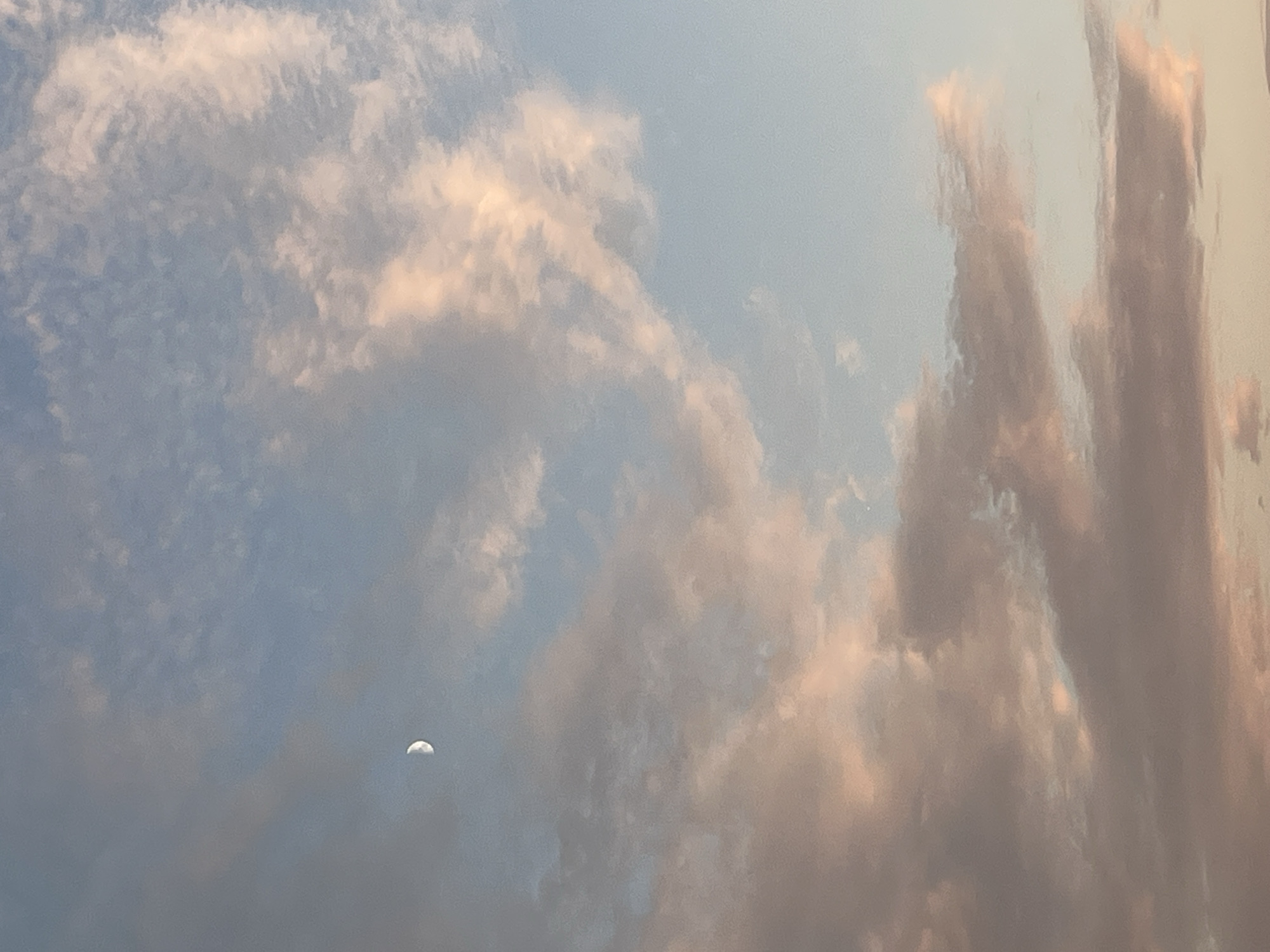 The image is a photograph of the sky with clouds. The sky is a pale blue, with scattered clouds that are tinged with pink and orange, suggesting it might be near sunrise or sunset. A sliver of the moon is visible in the upper left quadrant of the image. The image appears to have been taken from an outdoor location. There are no people, subjects, or objects in the foreground. The environment depicted is a sky with clouds and the moon. The image does not provide enough information to determine a specific location in the city of Ureki, Georgia.