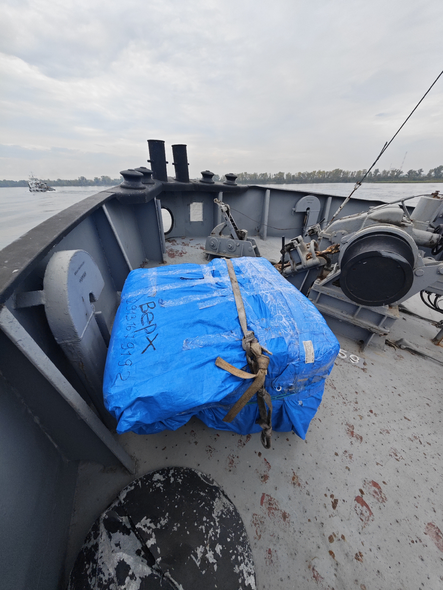 The image shows a bundle wrapped in blue plastic on the deck of a boat. The bundle is secured with straps and has some writing on it, including "BERX" and a series of numbers. The boat is on a body of water, likely a river, with trees and buildings visible in the background. The sky is overcast. The scene appears to be in the city of Ermolaevo, Russia, although no specific landmarks are identifiable to confirm this. The image is taken from a perspective looking forward on the boat. There are no people visible in the image. The deck of the boat is made of concrete and shows some wear and tear. There is also a piece of equipment on the deck, possibly a winch or anchor mechanism.
