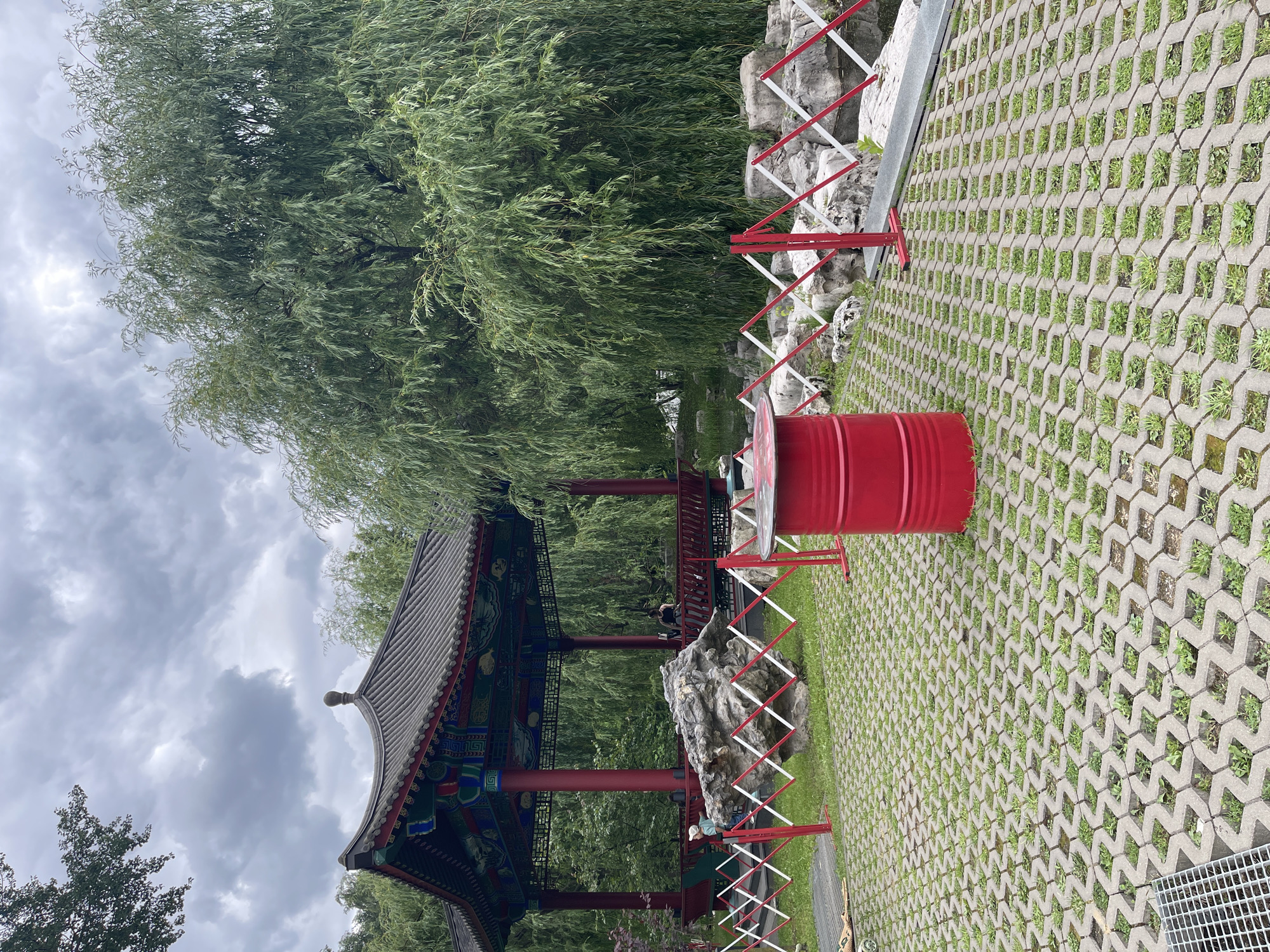 In an outdoor setting, a traditional Chinese-style pavilion with a dark, multi-tiered roof and vibrant red columns stands as the central subject. The pavilion is ornately decorated with colorful painted details. To the right of the pavilion, a large, weathered grey rock is situated. In front of the rock and extending across the image is a temporary barrier made of red and white crisscrossed metal supports. To the right of the barrier is a red cylindrical object, possibly a barrel or drum, resting on a ground surface composed of interlocking grey concrete pavers with patches of green grass growing in between. A large willow tree with flowing branches dominates the background, its leaves creating a lush green canopy. The sky above is overcast with grey clouds. There are several people visible within the scene; a few are standing on the walkway of the pavilion, and one person is seated near the base of the pavilion on the right side, partially obscured by the barrier. Another person is visible in the distance within the pavilion. The overall atmosphere appears calm and serene, with the natural elements of the tree and rocks contrasting with the man-made structures. The lighting suggests it is daytime, likely overcast or a cloudy day. No visible text is present in the image.