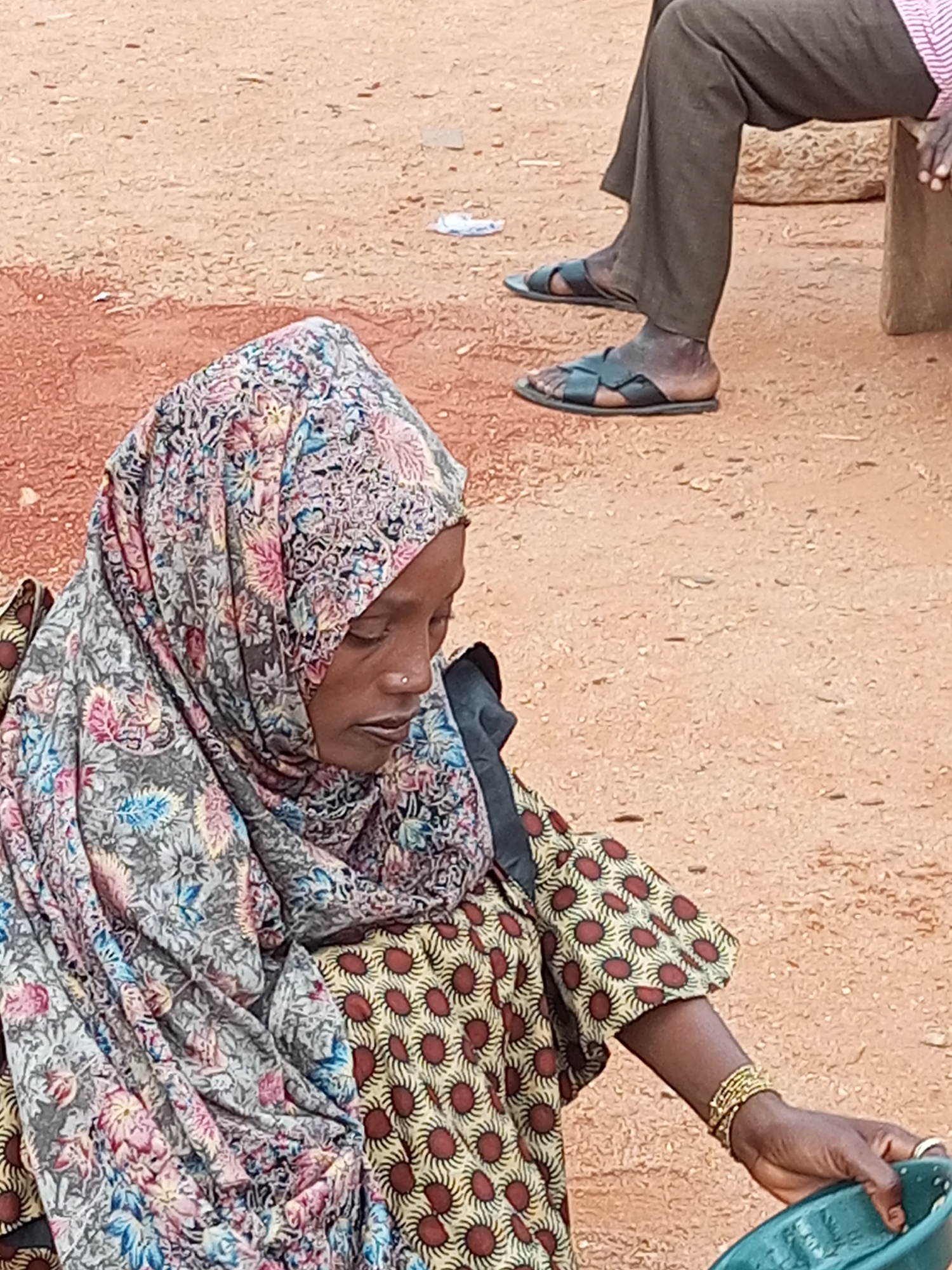 A woman wearing a colorful hijab and a patterned dress is seen from the waist up, sitting outdoors on sandy ground. She has a nose ring and appears to be looking down. Her right arm is visible, adorned with gold bracelets, and she is holding a green bowl. In the background, the lower legs and feet of another person wearing sandals and dark trousers are visible. The environment suggests a casual outdoor setting, possibly in a market or residential area. The scene is captured in Gombe, Nigeria.