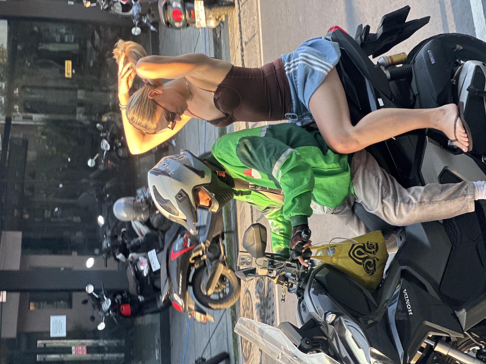 In a street scene in North Kuta, Indonesia, two individuals are positioned on a black motorcycle. A person wearing a green jacket and a helmet with a white and grey design is seated on the motorcycle, appearing to be the driver. They are holding the handlebars, and a yellow bag with a black emblem is visible on the left side of the motorcycle, near their legs.
Behind the driver, a second person, a woman with blonde hair, is leaning back against the motorcycle. She is wearing a brown top and denim shorts. Her hands are raised, with one hand touching her hair, and she is looking down. She is wearing sunglasses and a bracelet. Her right foot is resting on the motorcycle, wearing a flip-flop.
The motorcycle appears to be parked or stopped on a paved surface, possibly a street or sidewalk, with other motorcycles visible in the background, suggesting an urban environment. The lighting suggests it might be late afternoon or early evening, with a warm, golden hue from the sun. The overall impression is a casual moment captured while on or near a motorcycle in a busy area. There is visible text on the motorcycle: "HONDA".