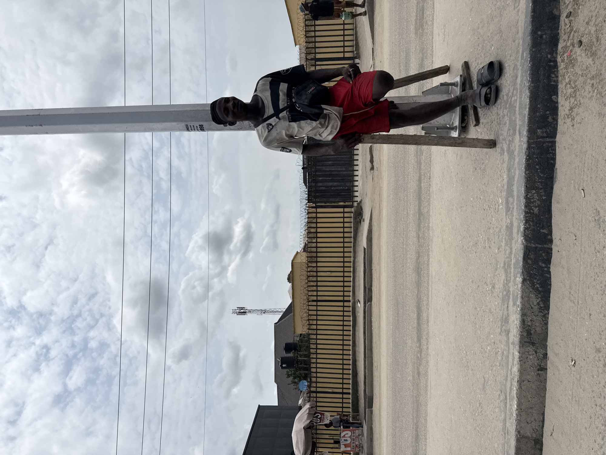 The image depicts an outdoor street scene in Ado, Nigeria, captured during the daytime under a bright, cloudy sky.
The main subject is a young man, appearing dark-skinned, who is standing near the edge of a paved road. He leans against a tall, light-gray utility pole, which is reinforced at its base with wooden planks. He is dressed in a white short-sleeved shirt with black horizontal stripes, red shorts, and dark slippers. A black fanny pack or crossbody bag, visible across his chest, has the words "CHIMU" and "ABY" printed on it. He looks towards the camera with a neutral expression.
In the background, behind a black metal fence topped with barbed wire, are various buildings, including one with multiple black water tanks on its roof. A communications tower can be seen further in the distance. The scene includes a paved road and a concrete curb. A few other blurry figures are present in the deeper background.
Notable details include text on the utility pole: "BUSINESS," "0805 060 1620," and "0803 709 0398." Near the bottom right, next to what appears to be a small white tent or umbrella, partially visible signs include the phrase "WE BUY YOU BRAINS" and "ST". The overall image conveys a slice of daily life in a bustling urban or semi-urban environment.