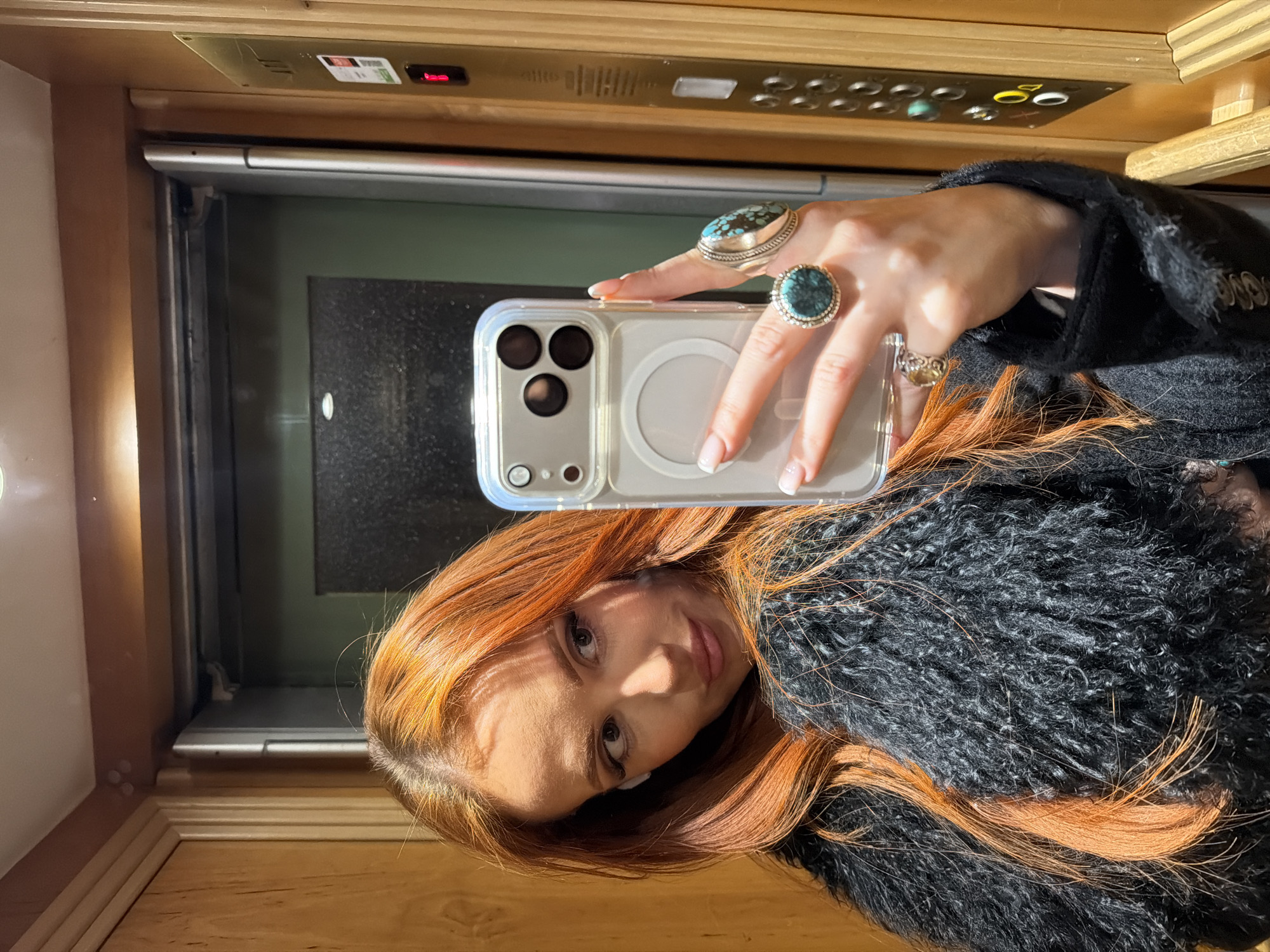 The image displays a woman taking a mirror selfie inside an elevator. She possesses long, wavy, reddish-orange hair and is dressed in a dark, shaggy or textured coat. White wireless earbuds are visible in her ears. Her right hand holds a light-colored iPhone, encased in clear plastic with a visible MagSafe ring, angled towards the reflection. This hand exhibits manicured nails with white tips and is adorned with multiple silver rings, notably two large ones featuring prominent turquoise stones on her middle and ring fingers. The woman's gaze is directed towards her right, away from the phone's camera, with a subtle expression.
The elevator interior is characterized by wood-paneled walls. To the woman's right, a brass-colored control panel is affixed, displaying a red digital '1' at its top, indicating the current floor. Below this digit are several silver buttons and various stickers, including a distinct green one. A dark, metallic elevator door is partially visible in the reflection behind the woman. The scene is set within an elevator in Paris, France.
