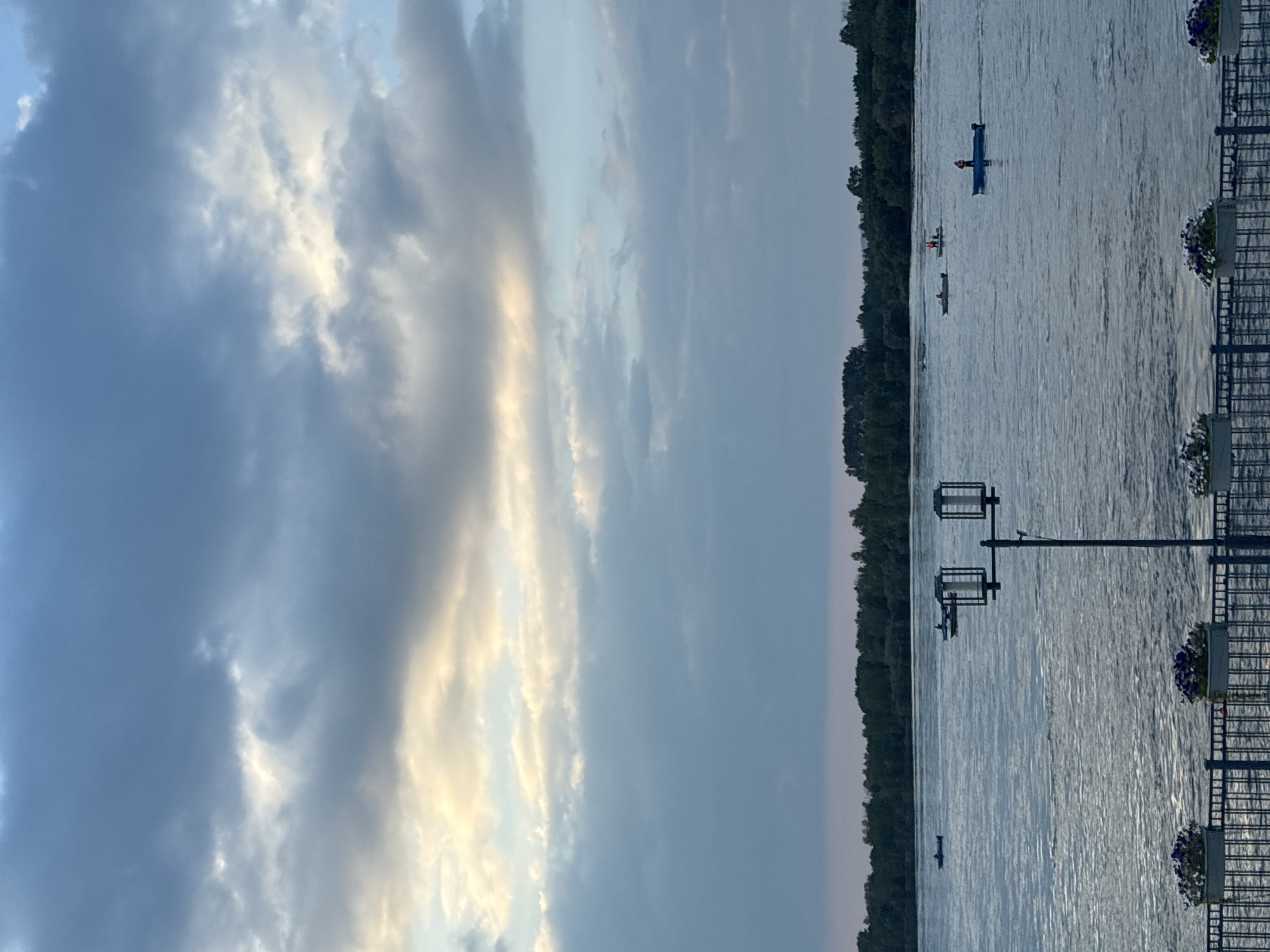 The image shows a wide river with several small boats on it. The boats appear to be occupied by people, some of whom might be fishing. The riverbanks are lined with trees. In the foreground, there is a fence with planters containing flowers. A lamppost is also visible in the foreground. The sky is cloudy with some patches of light breaking through. The overall impression is a serene, natural scene, likely during the late afternoon or early evening.
This scene could be located on the Angara River in Irkutsk, Russia. The Angara River is a significant waterway that flows through Irkutsk, and it is common for people to engage in recreational activities such as boating and fishing on the river. The presence of trees along the bank and the general landscape are consistent with the scenery found in Irkutsk.