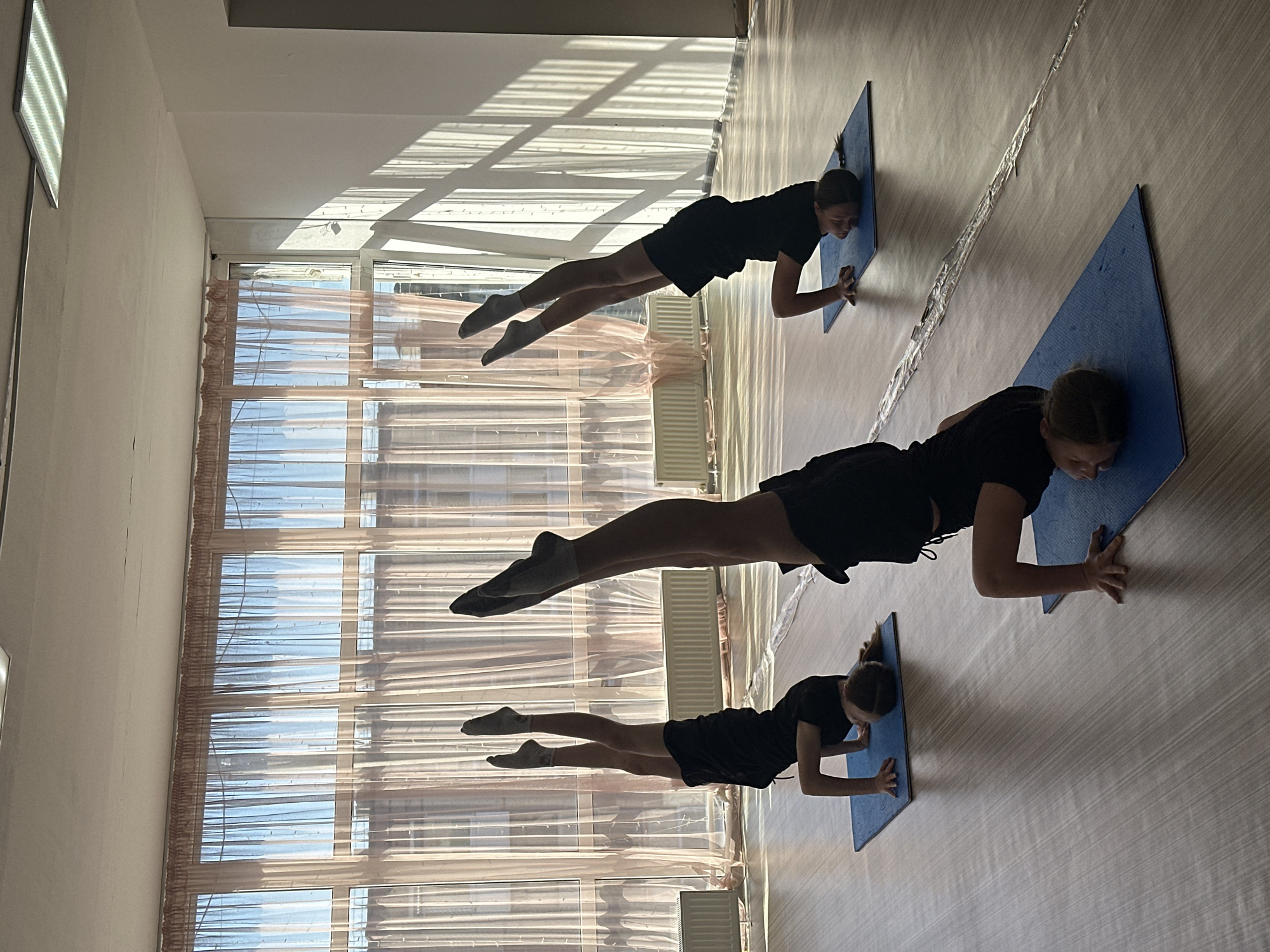 Three individuals are engaged in a plank exercise, each on a blue exercise mat, within a bright, indoor space. The floor appears to be made of light-colored linoleum, and light streams through a large window covered with sheer curtains. The location is potentially in Votkinsk, Russia, as indicated by the user's instruction.