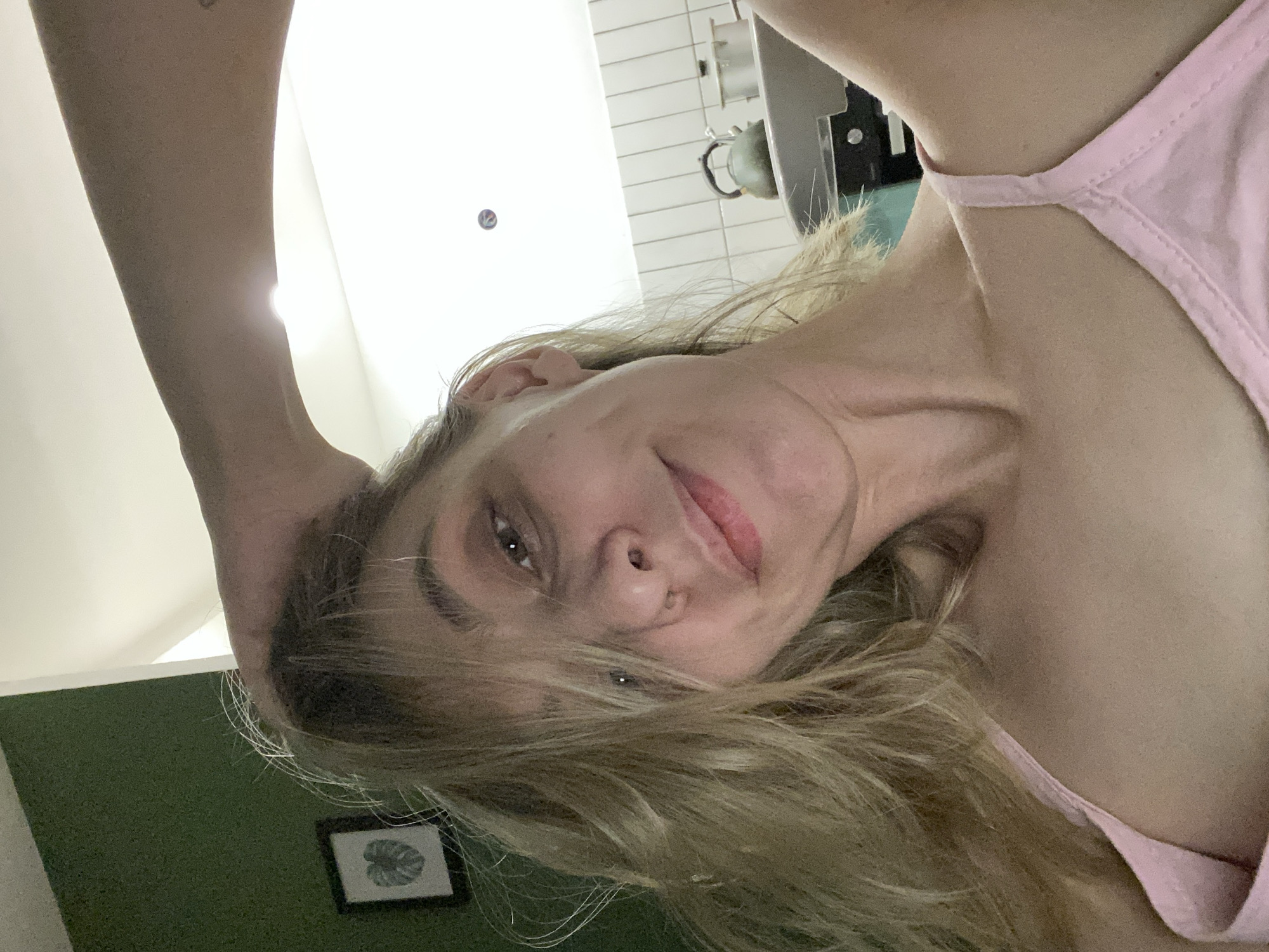 A close-up selfie depicts a female subject with blonde hair, fair complexion, and brown eyes, facing forward with a subtle smile. She is wearing a pink spaghetti-strap top. Her right forearm is raised, and her hand is positioned in her hair above her head. The background on the left features a dark green wall with a black-framed picture containing a white Monstera leaf silhouette. The background on the right shows a kitchen area with white subway tiles, a visible metal teapot, and part of a metal pot. The specific location within Chaikovsky, Russia, cannot be determined from the visual evidence.