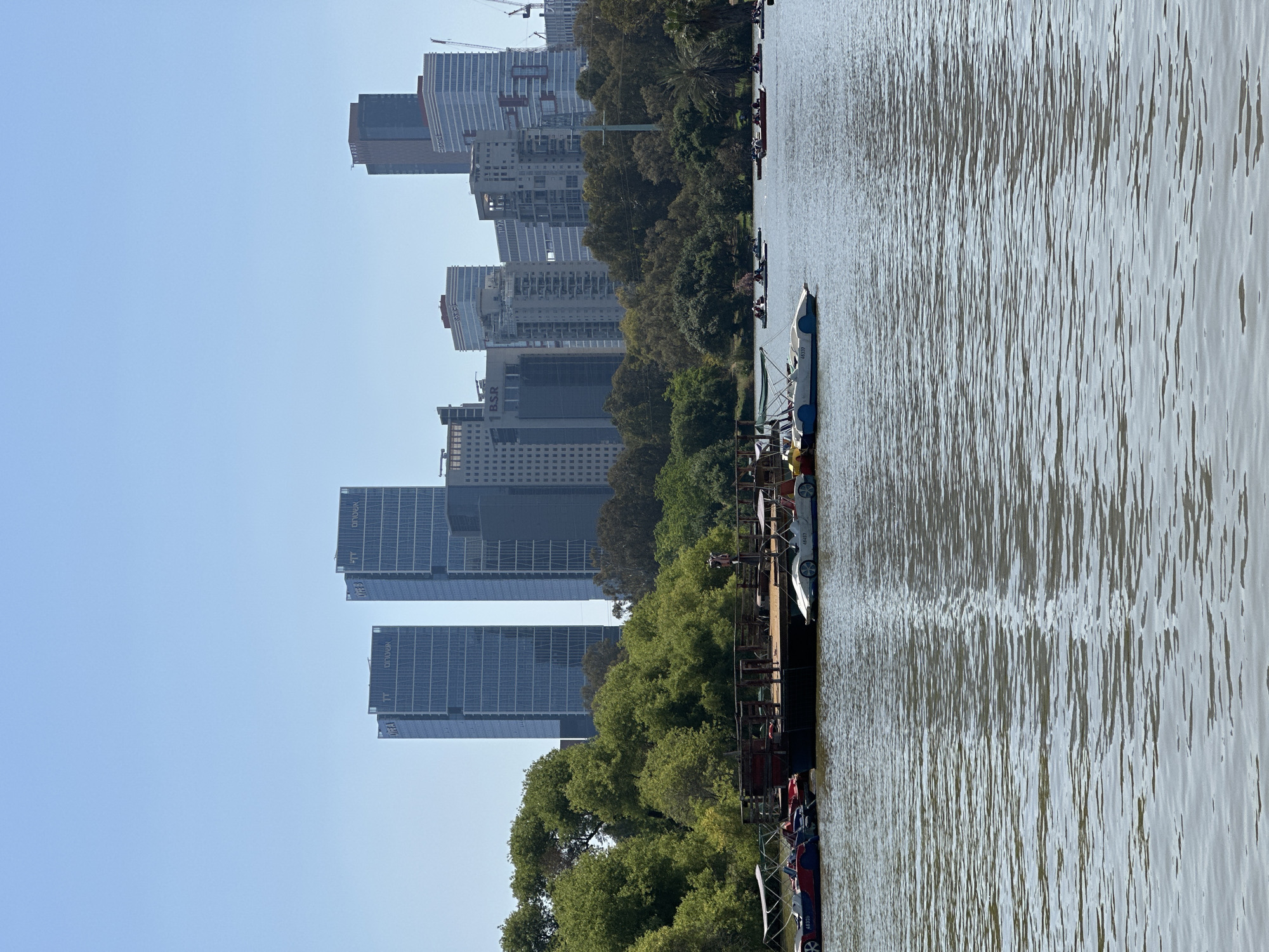 The image shows a waterway in Tel Aviv, Israel, likely the Yarkon River. On the left, the skyline of the city is visible, with several modern high-rise buildings. Between the water and the buildings is a line of trees and green vegetation. On the right is the water, which appears calm with subtle ripples. A pier or dock extends into the water, and there are several small boats or paddleboats docked there. There are people on the dock or nearby, but they are not clear enough to identify any specific actions or interactions. The sky is clear and blue.