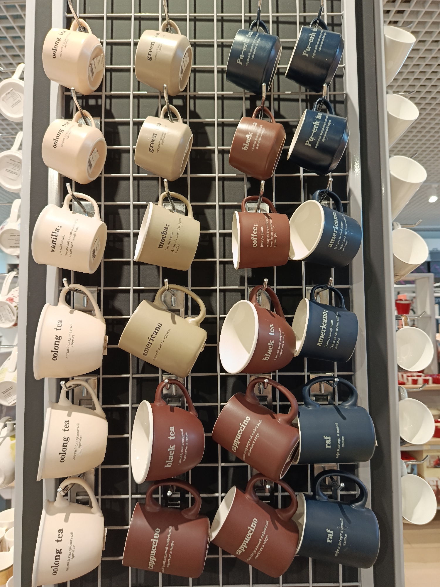 A display of novelty mugs, each hanging on a metal grid, is present in what appears to be a store in Moscow, Russia. The mugs are various colors and labeled with different drink names and descriptions in both English and Russian: "oolong tea", "vanilla", "cappuccino", "black tea", "americano", "mocha", "coffee", "green", "raf", "Pu-erh tea". The mugs are arrayed in rows, with each row consisting of a light tan mug, a tan mug, a brown mug, and a blue mug. The interior of each mug is white. Other similar mugs are visible in the background, indicating the display is part of a larger section. The scene is well-lit.