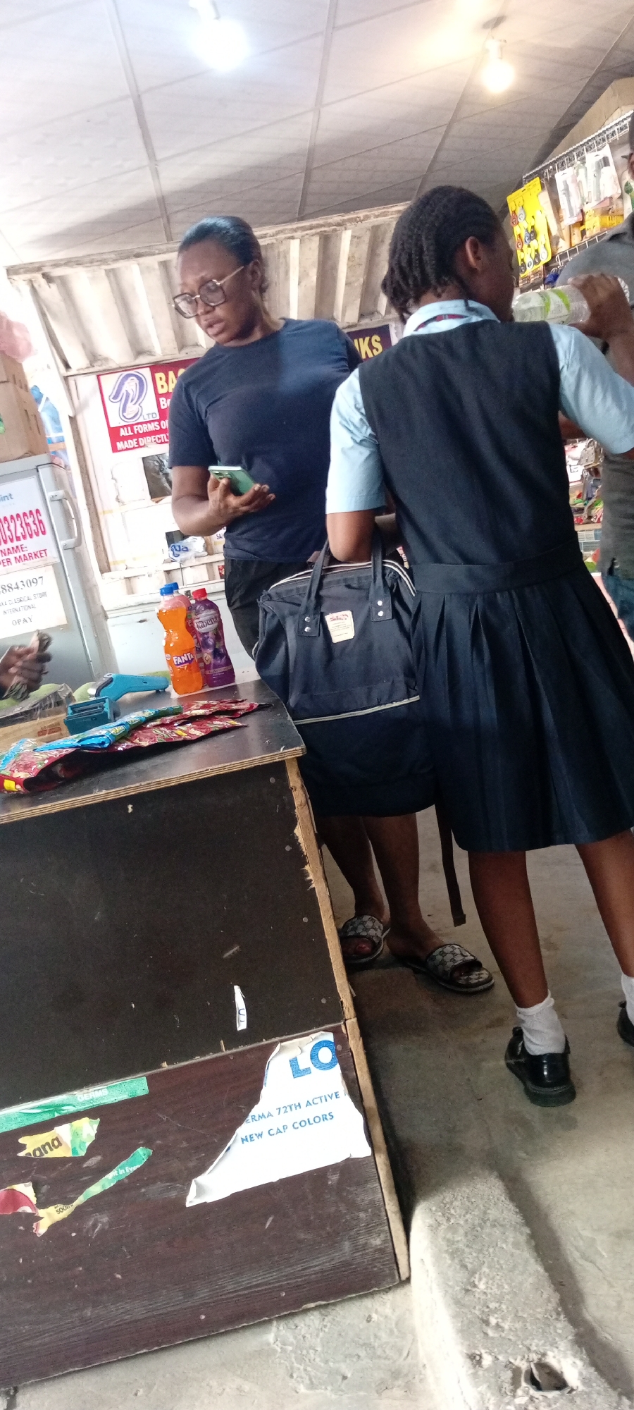 This media file shows two people, likely a woman and a young girl, standing at a counter in what appears to be a market or a small shop. The woman, wearing glasses and a dark blue t-shirt, is holding a smartphone. The girl is dressed in a school uniform consisting of a light blue shirt and a dark blue pinafore with a pleated skirt. She is also carrying a dark blue bag.
On the counter, several items are visible, including two bottles of drinks, one orange and one purple, and some colorful snack packages. A blue-colored receipt printer or scanner is also present. The background suggests an indoor setting with a metal roof and some signage. One sign, partially visible, has text that includes "BA" and "KS". Another sign, on the left, displays numbers and text that seems to be "SUPER MARKET". There are also torn pieces of paper with text, including "LO", "ERMA 72TH ACTIVE", and "NEW CAP COLORS".
The overall impression is a casual scene in a marketplace. The lighting suggests it could be daytime, and there are no strong indicators of specific weather conditions. The interaction between the individuals is not clear, but the presence of shopping items and a transaction device implies a purchase is either happening or about to happen. The setting appears to be in Nigeria, specifically within Aviation Quarters.