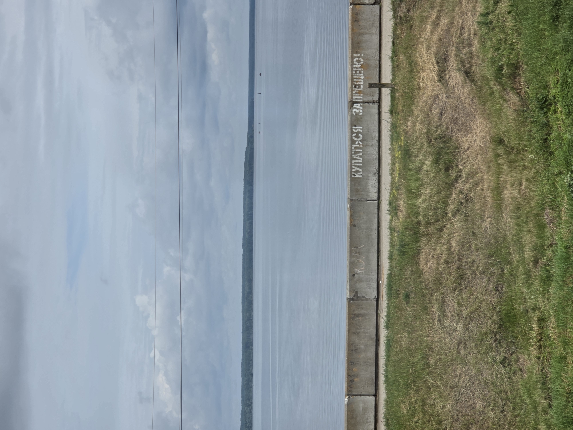 The image depicts a concrete barrier along the embankment of a wide body of water, likely a river or lake, under an overcast sky. The barrier, composed of several concrete blocks, is marked with white spray-painted Cyrillic text that reads "КУПАТЬСЯ ЗАПРЕЩЕНО!" which translates to "SWIMMING IS PROHIBITED!". In the distance, a treeline forms the horizon across the water. A few small boats or buoys are visible on the water's surface. In the foreground, a strip of dry and green grass is present. The scene appears to be located in Kozmodemyansk, Russia, based on the context provided.