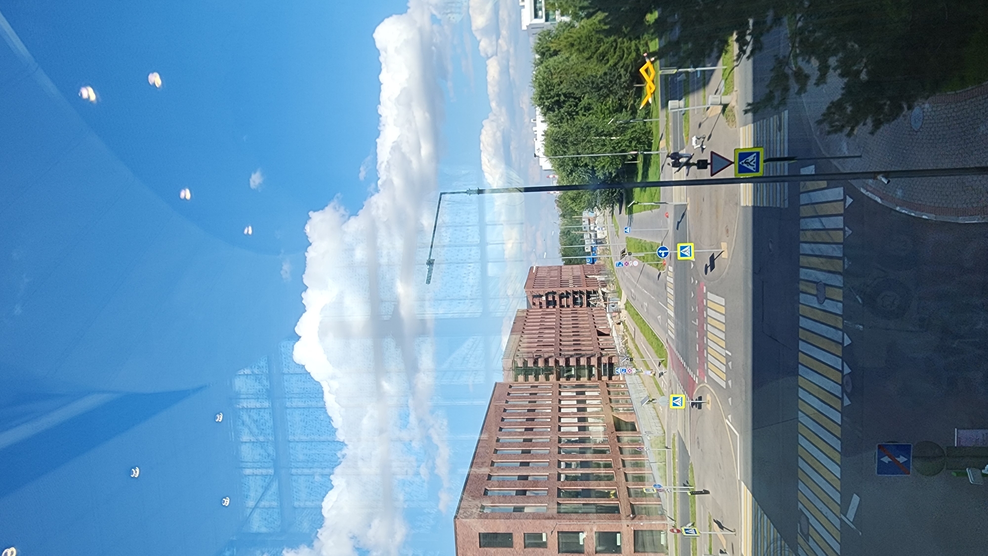 This is a photo taken from a high vantage point, likely an upper floor window, overlooking a modern urban intersection in Moscow, Russia. The view is dominated by a clear blue sky with scattered white clouds. Reflections of interior lights and the building's structure are visible on the glass.
In the foreground, a wide pedestrian crossing with yellow and white striped markings is prominent. Further into the scene, a street curves to the left, with an intersection featuring multiple road markings, including crosswalks and traffic lanes. Several yellow triangular road signs with black pictograms, likely indicating pedestrian crossings or caution, are visible.
On the left side of the street, a long, multi-story building with a facade of red brick and numerous windows stands. Adjacent to it, another similar building is under construction or renovation, with visible scaffolding and exposed structural elements.
A street light with a single lamp extends over the intersection. Beyond the buildings and across the street, lush green trees form a dense line. In the distance, other modern buildings and some construction cranes are visible.
Two people and a small dog are walking on a sidewalk near the intersection. The overall impression is a clean, well-maintained, and modern urban environment under a bright, sunny sky.